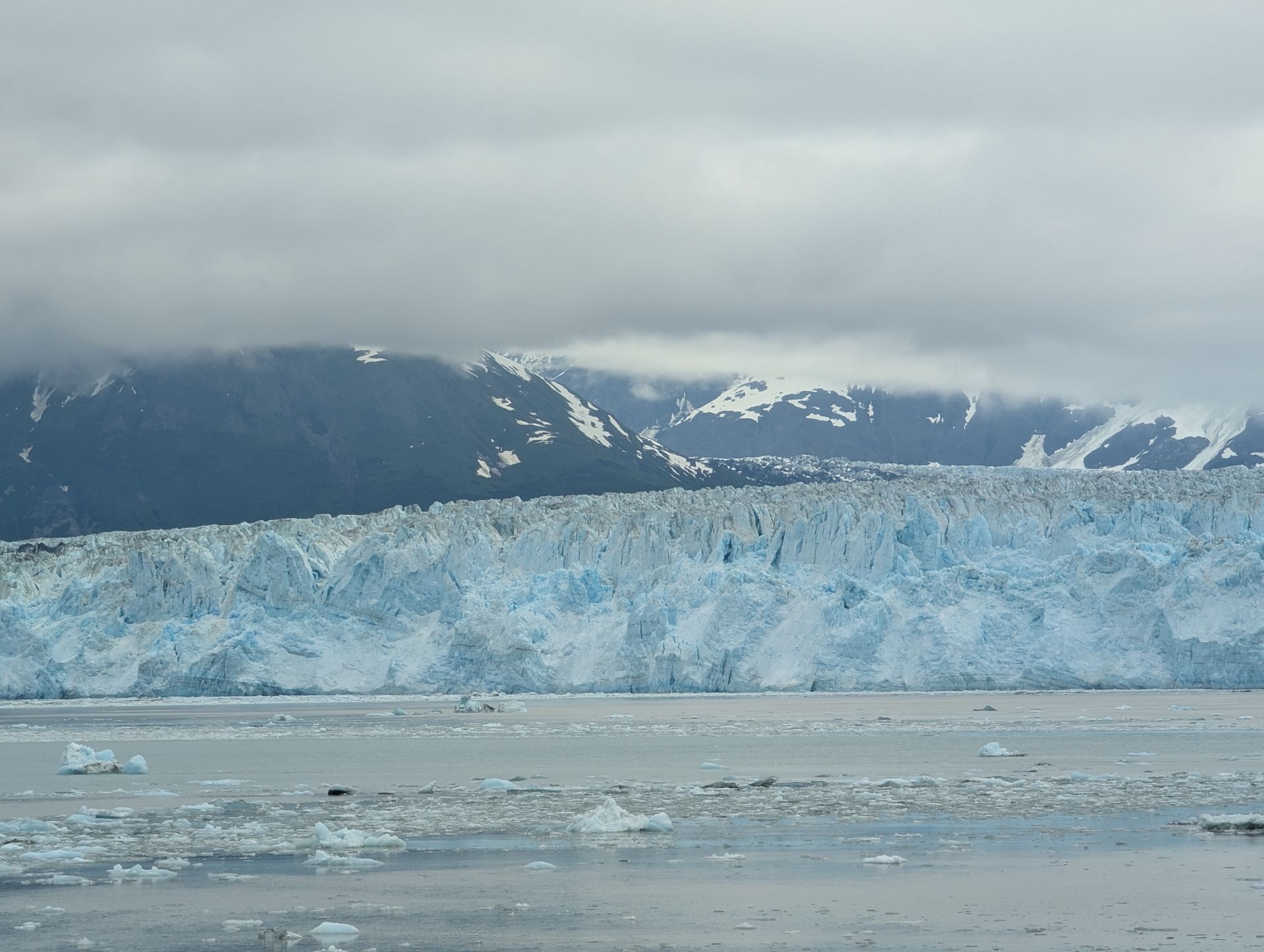 A vast, blue glacier front meets the ocean, with icy debris scattered. Mountains and cloudy skies are in the background.