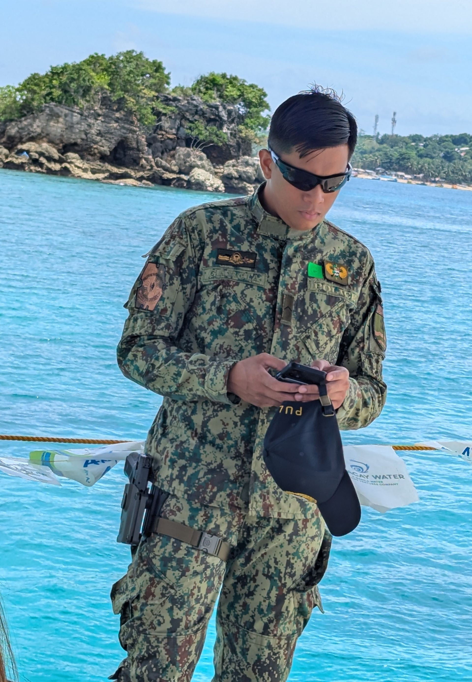 A uniformed officer with sunglasses holds a hat, looking down. Outdoors on a boat, the ocean and an island are in the background.