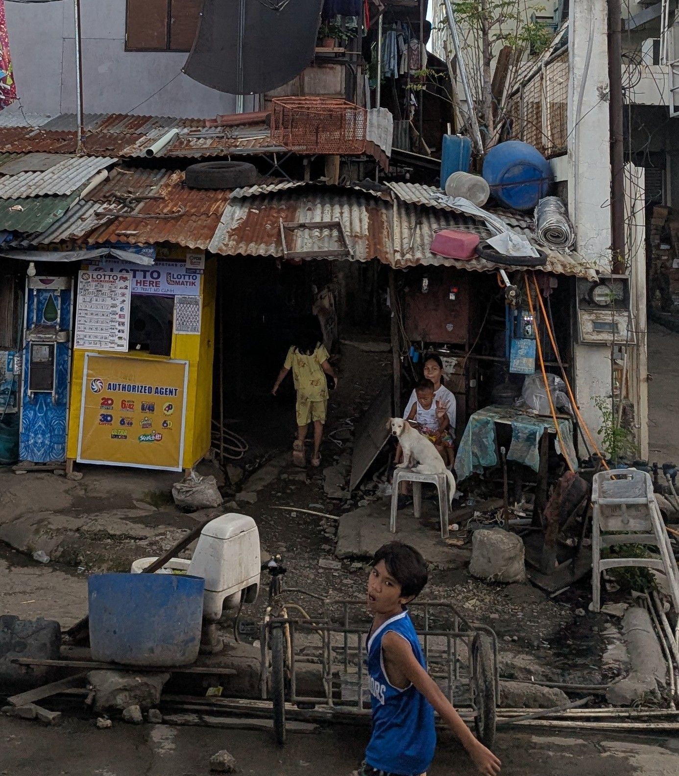 A child runs past a shop with corrugated metal roof. Another child and woman are inside.