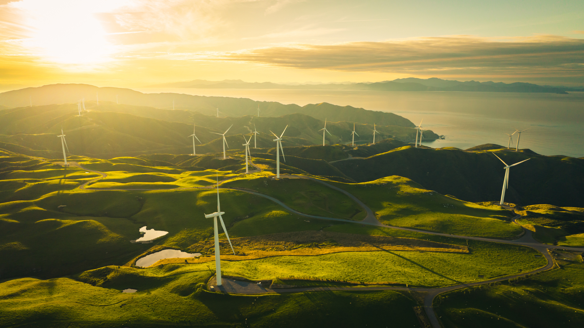 An aerial view of a wind farm on a hill overlooking the ocean at sunset.