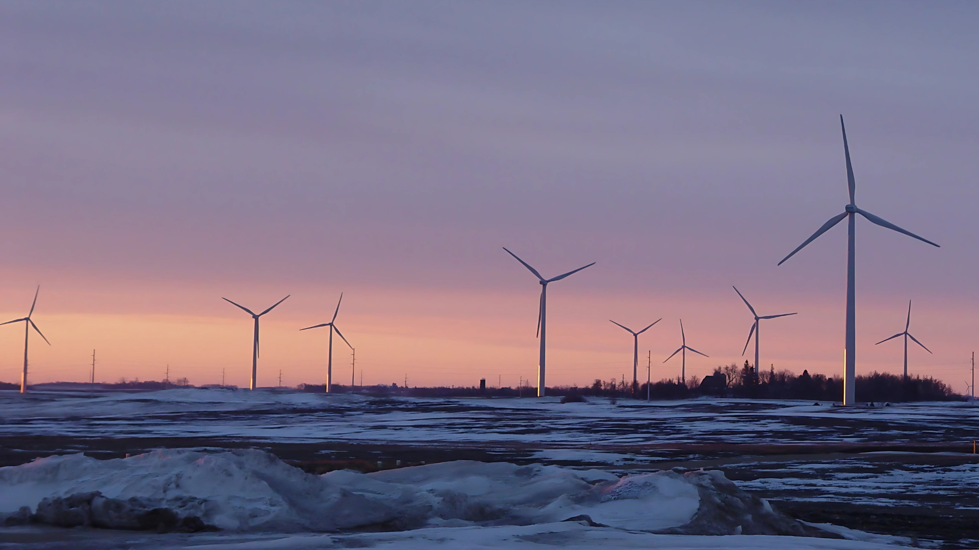 A row of wind turbines in a snowy field at sunset