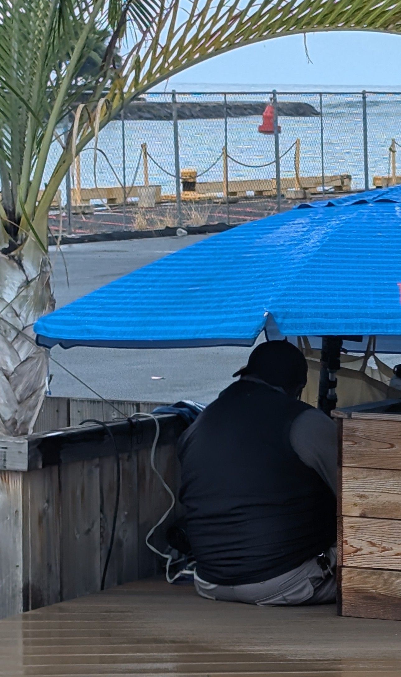 A man is sitting under a blue umbrella in front of a body of water.