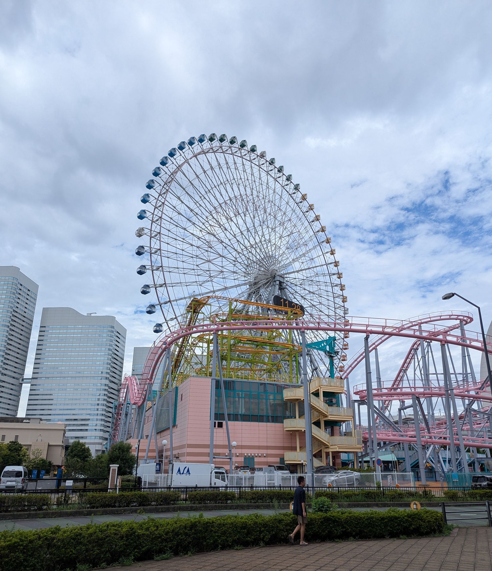 Ferris wheel and roller coaster at a park in front of buildings, cloudy sky.