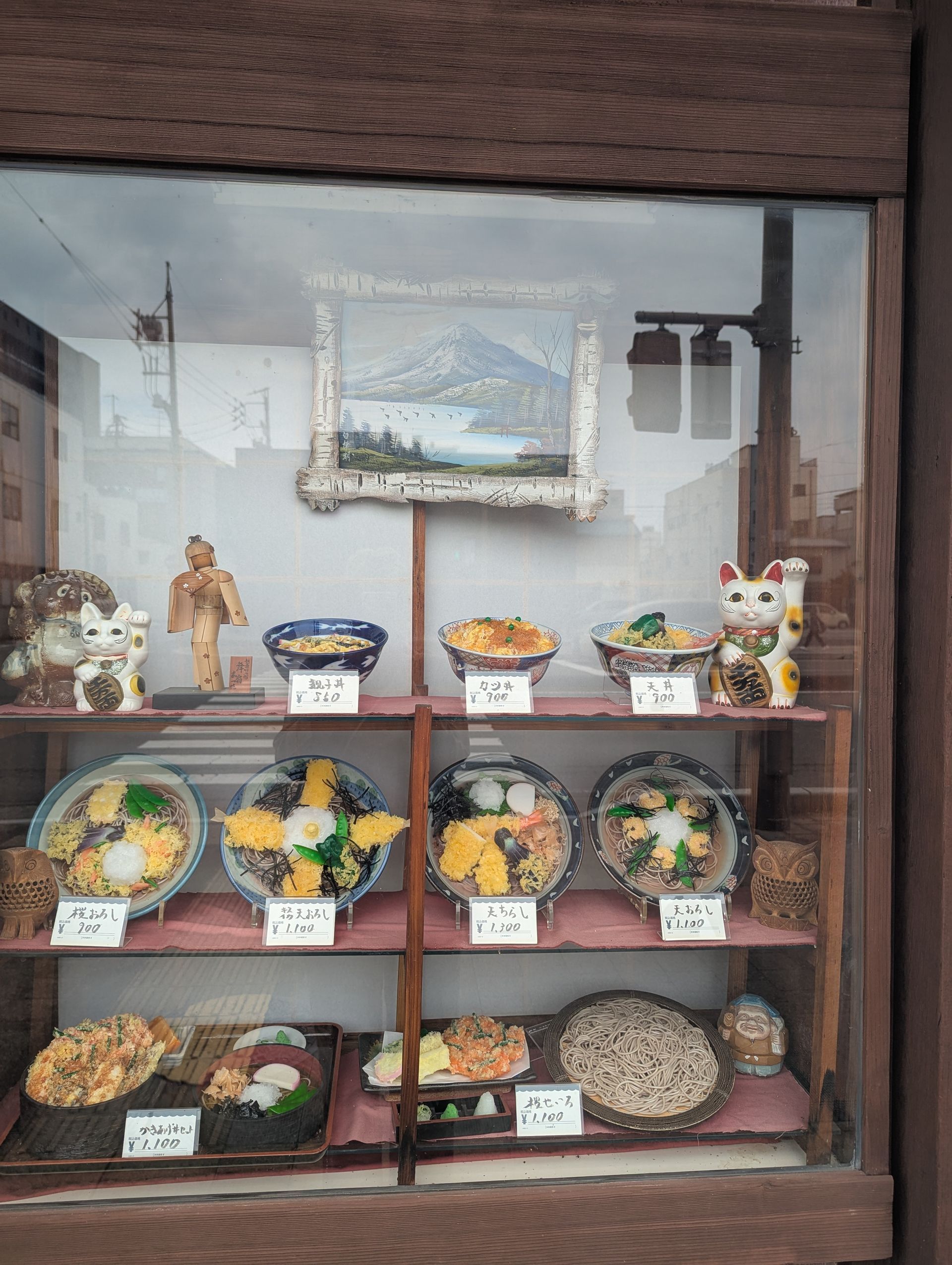 A restaurant display window with various colorful, appetizing food models. A painting and lucky cat statue are also visible.