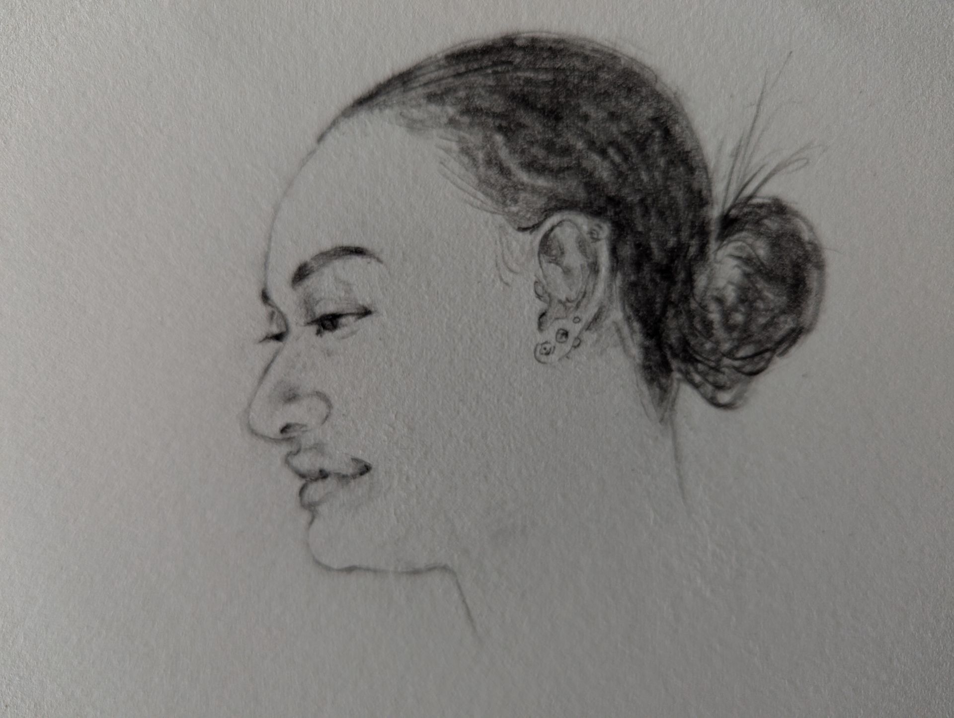 Profile view of a person with a bun hairstyle, drawn in pencil, showing facial features and ear piercings.
