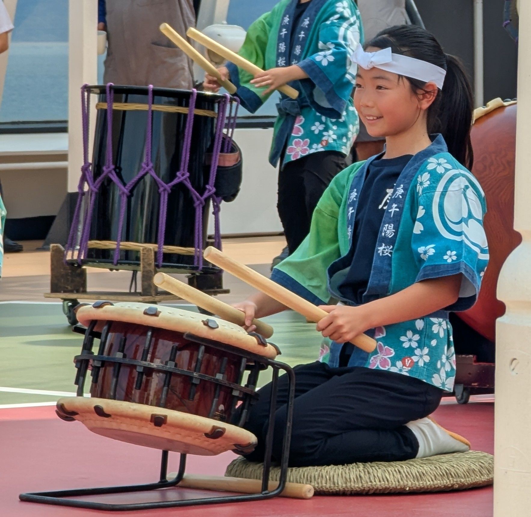 Young girl in teal kimono plays a Japanese drum outside, smiling.