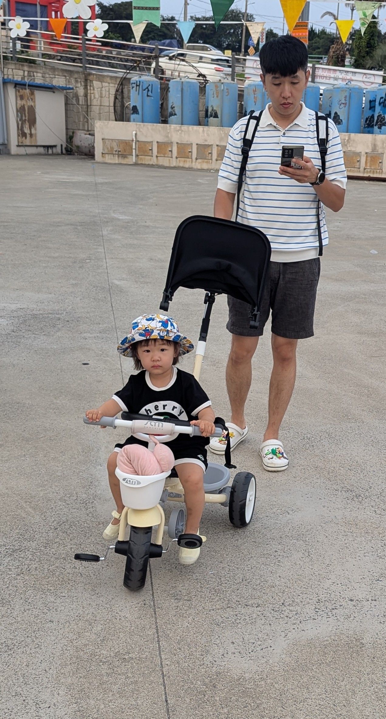 A child riding a tricycle is pushed by a man looking at his phone on a paved surface.