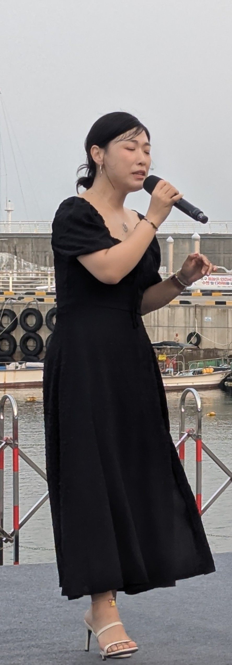 Woman singing into a microphone on a stage. She wears a black dress and heels, with a harbor in the background.