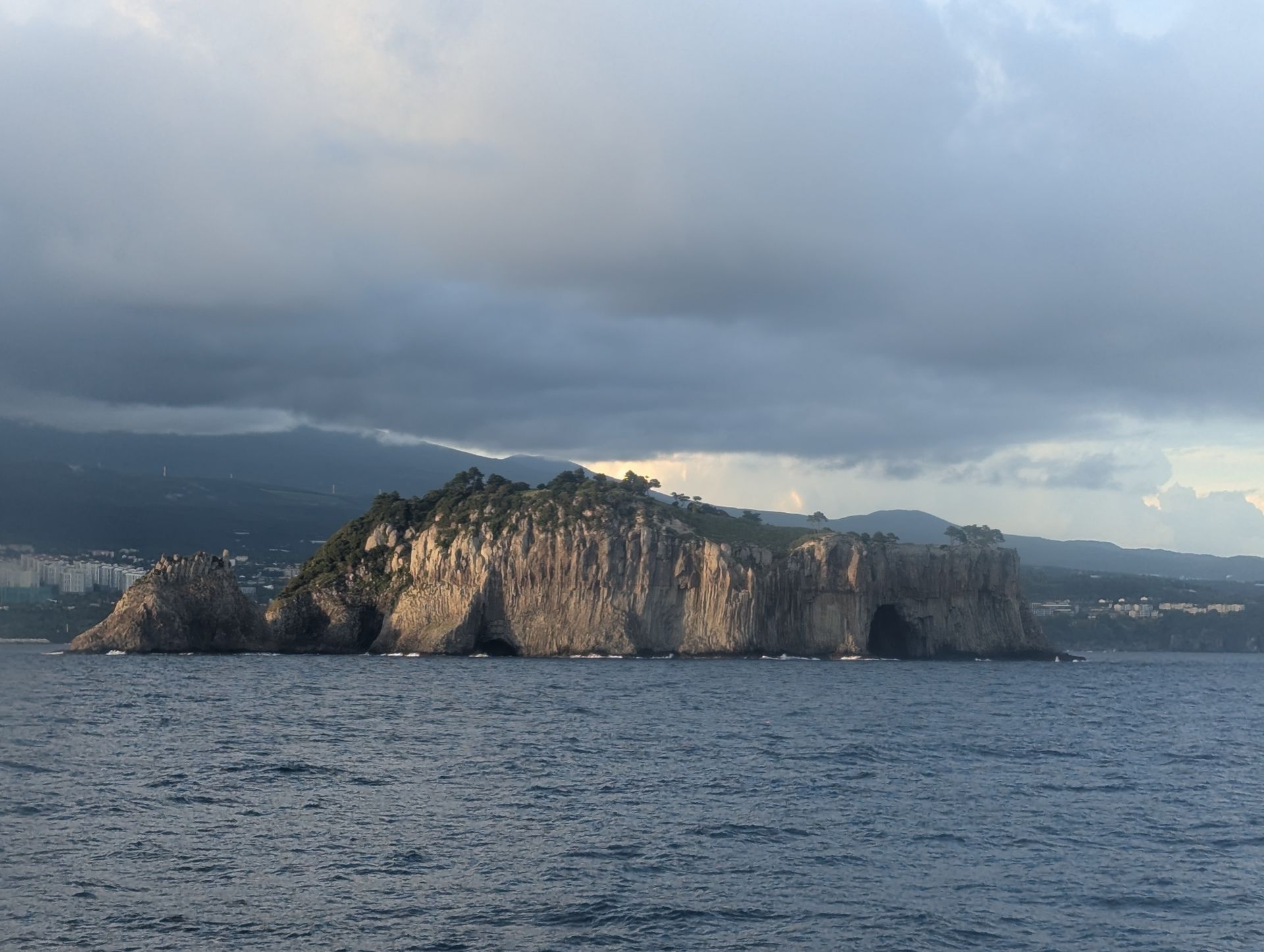 Island with rocky cliffs and lush vegetation, partially illuminated under cloudy skies.
