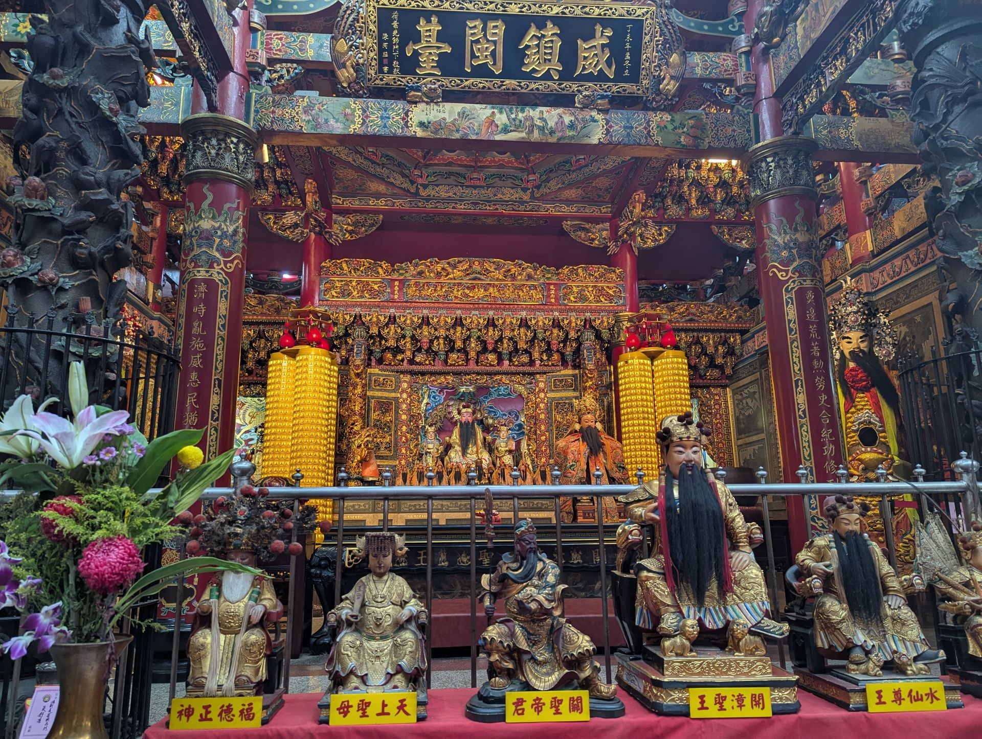 Ornate gold and red Taiwanese temple interior with statues, altar, and floral arrangements.