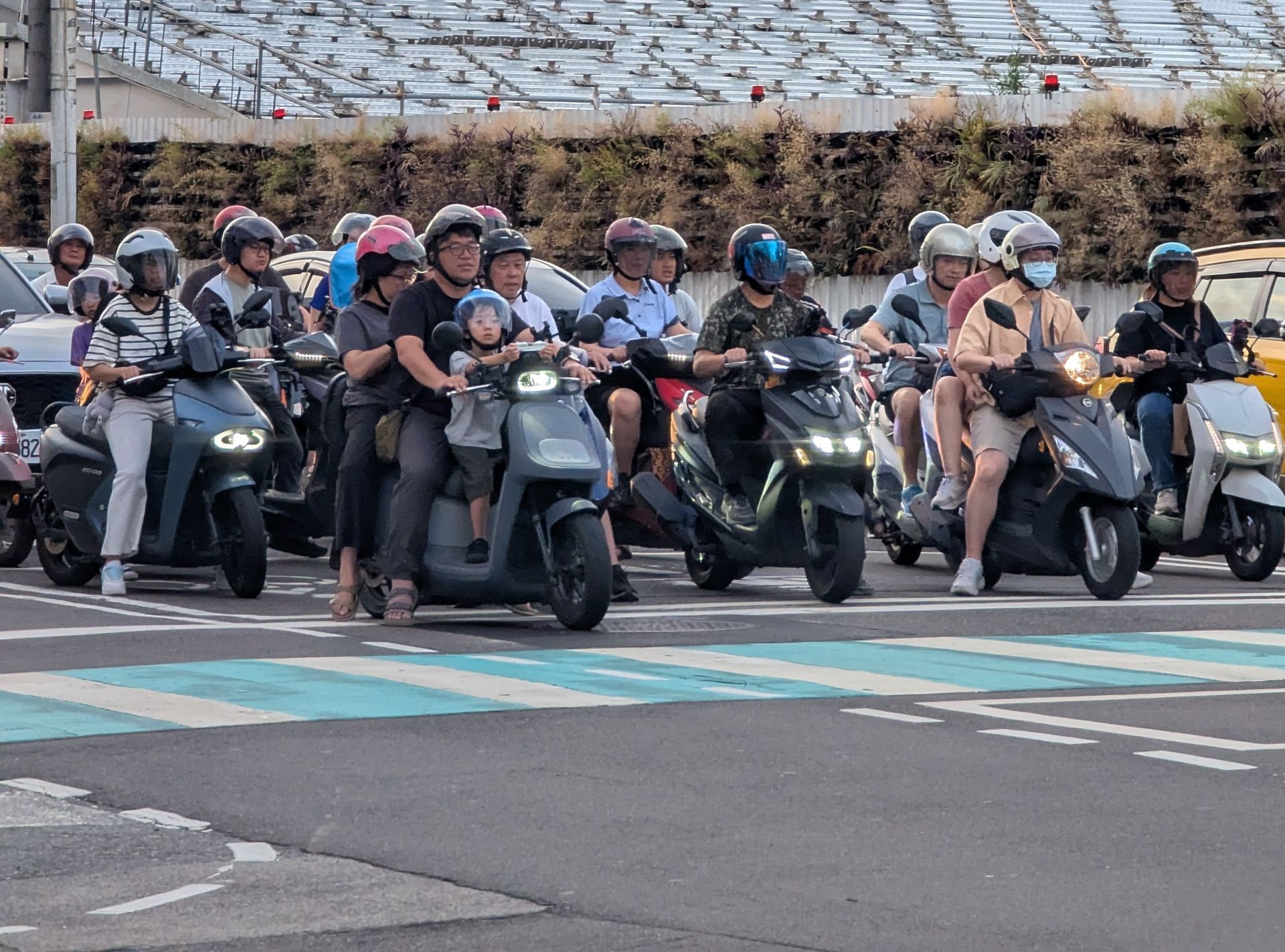 Motorcycles with riders at a crosswalk in Taiwan. People wearing helmets. Grey and black scooters.