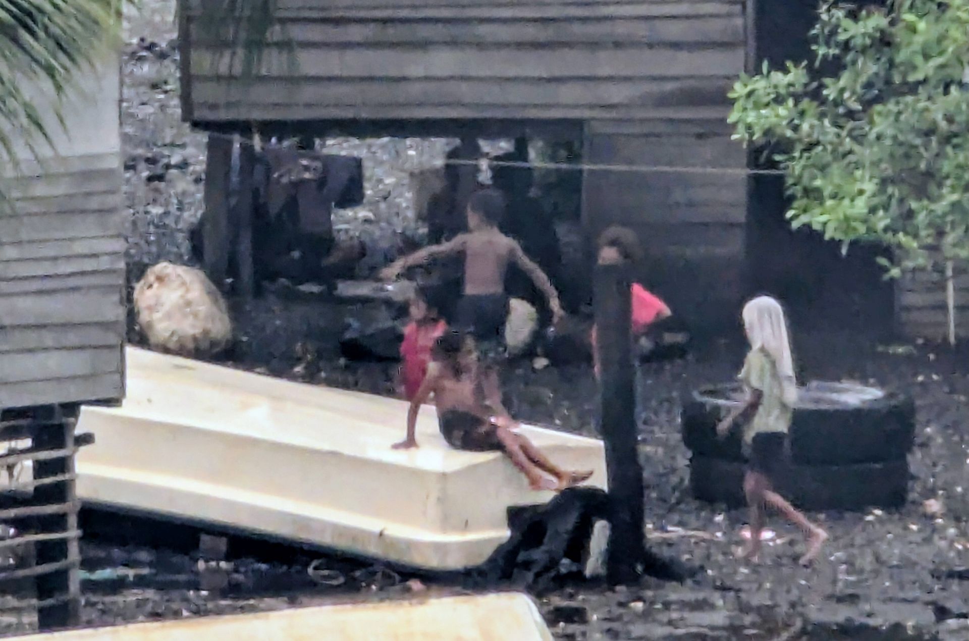 Children playing in the rain near a makeshift slide and tires in a muddy, outdoor environment.