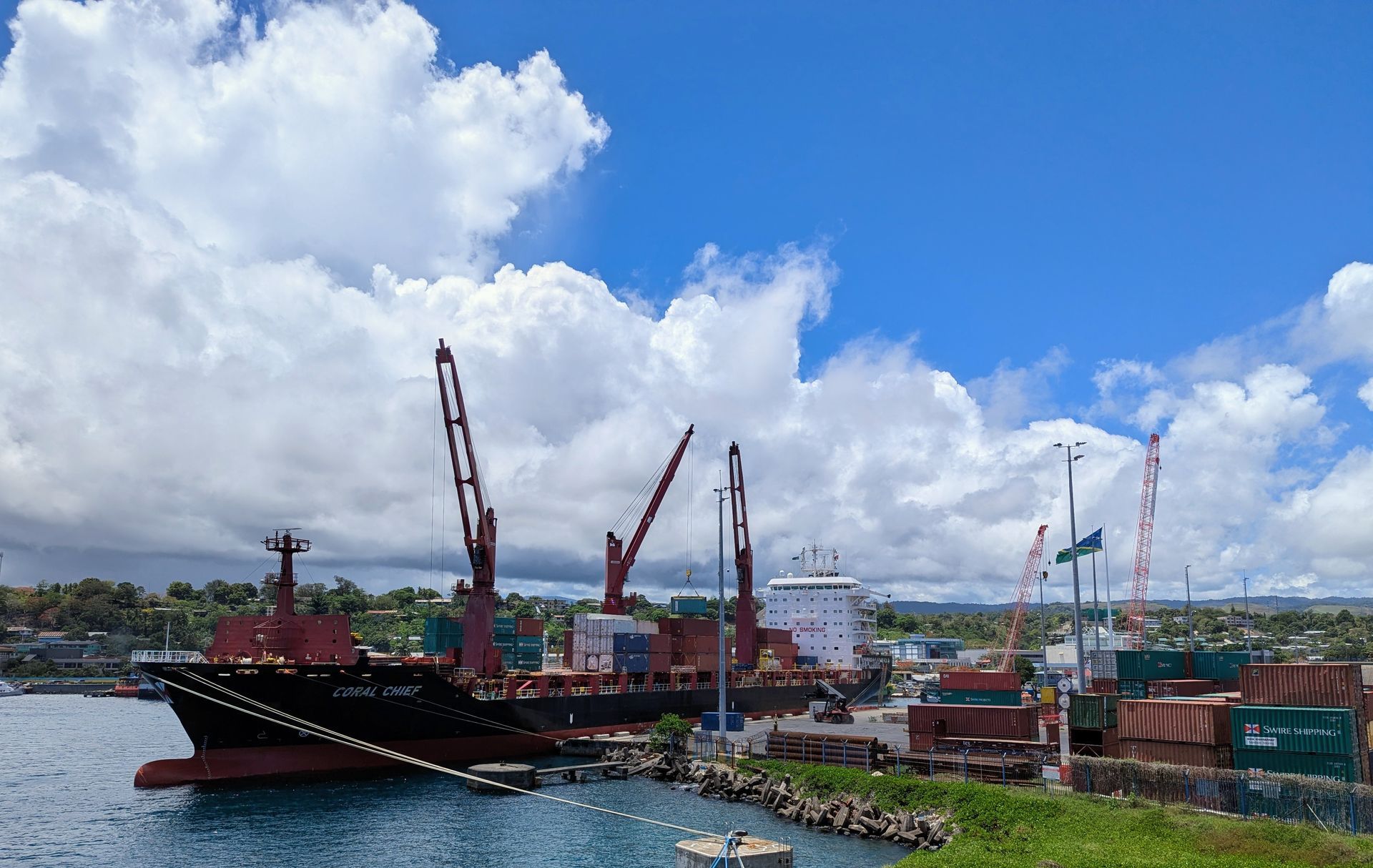 Cargo ship docked at harbor under a blue sky with fluffy white clouds; cranes and shipping containers visible.