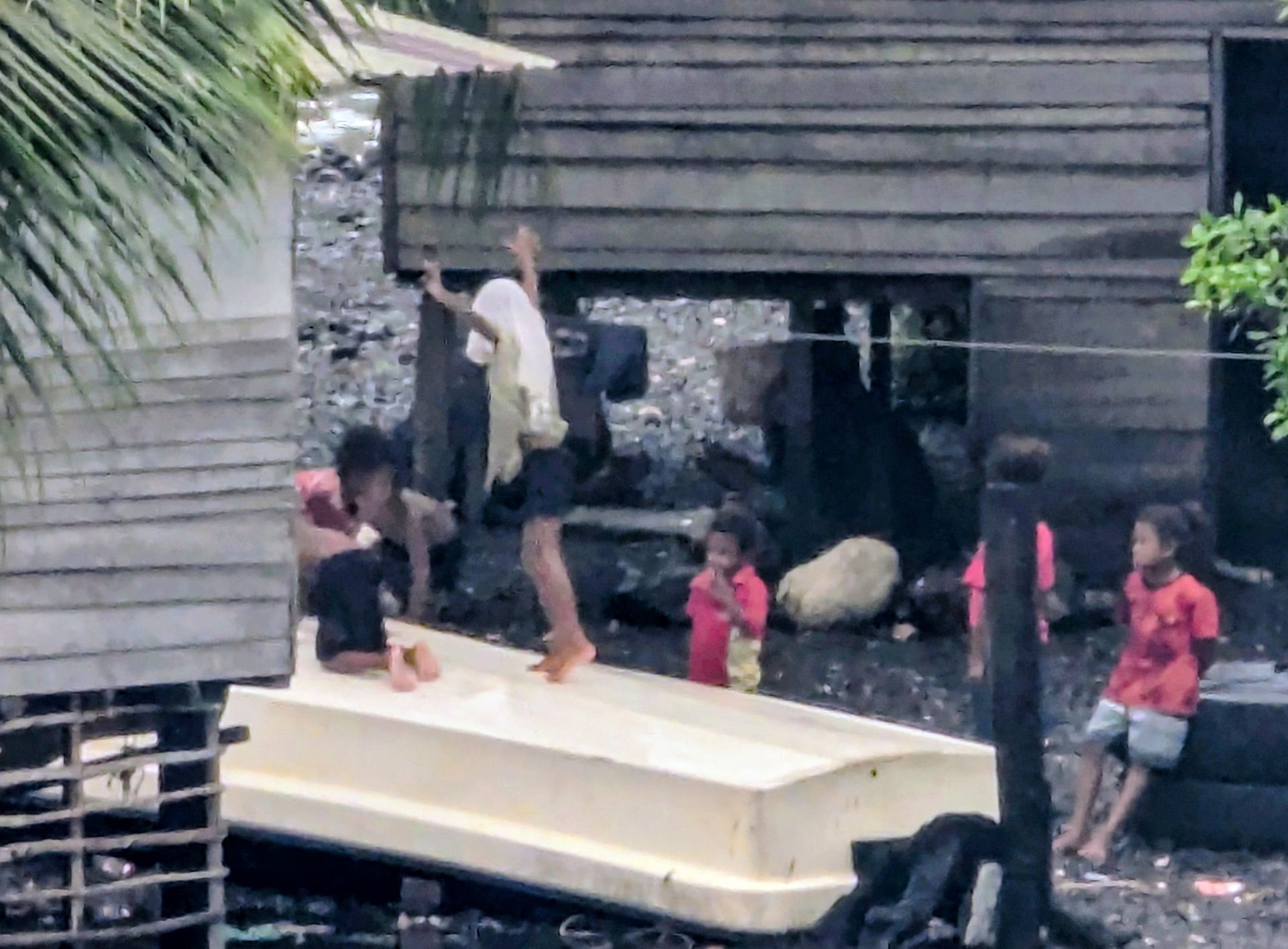Children playing near a boat, next to a wooden building. One child jumps, others sit or stand.