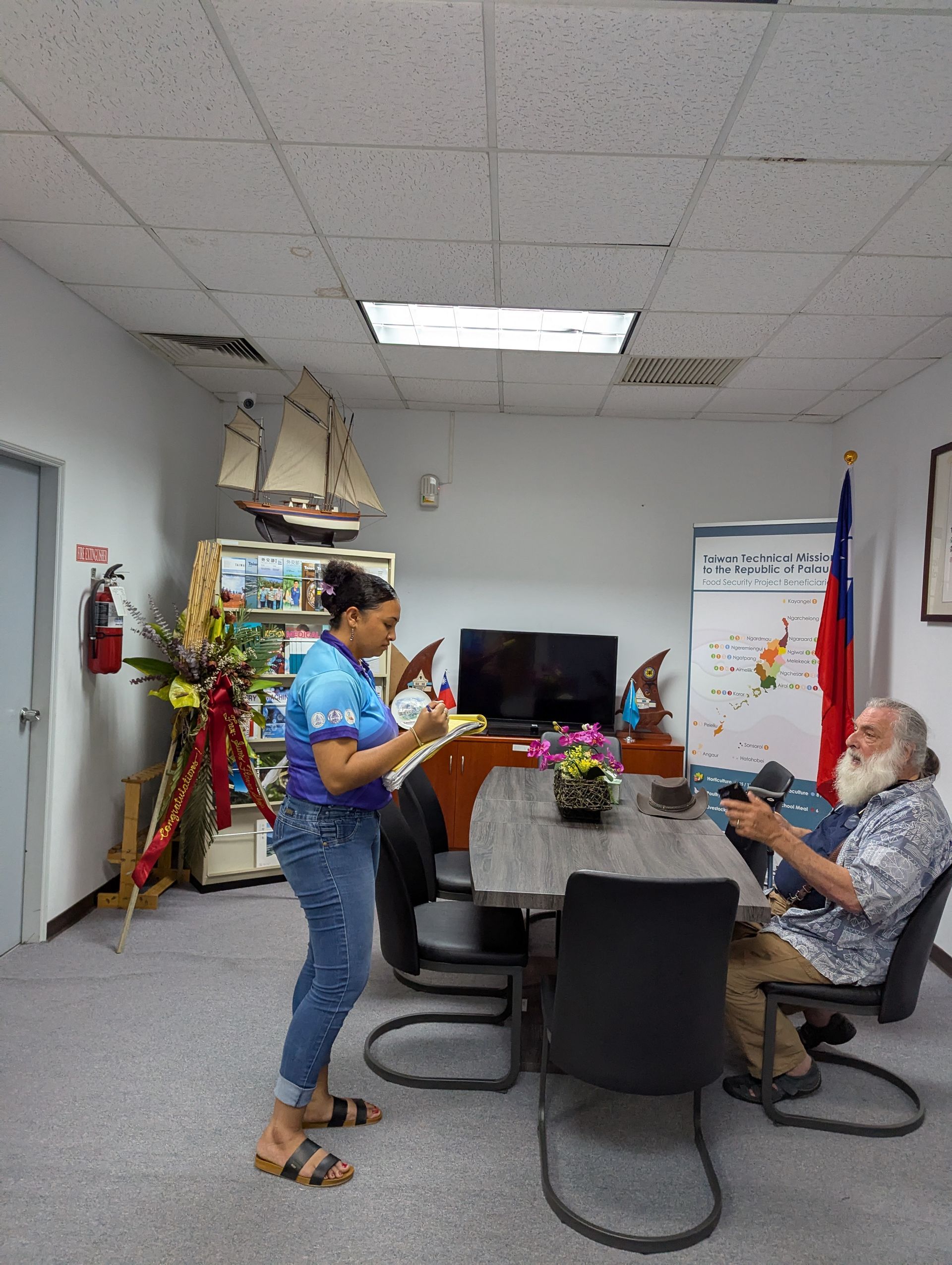 Woman and man in an office. Woman stands, man sits. A sailing ship model and a Philippine flag are visible.