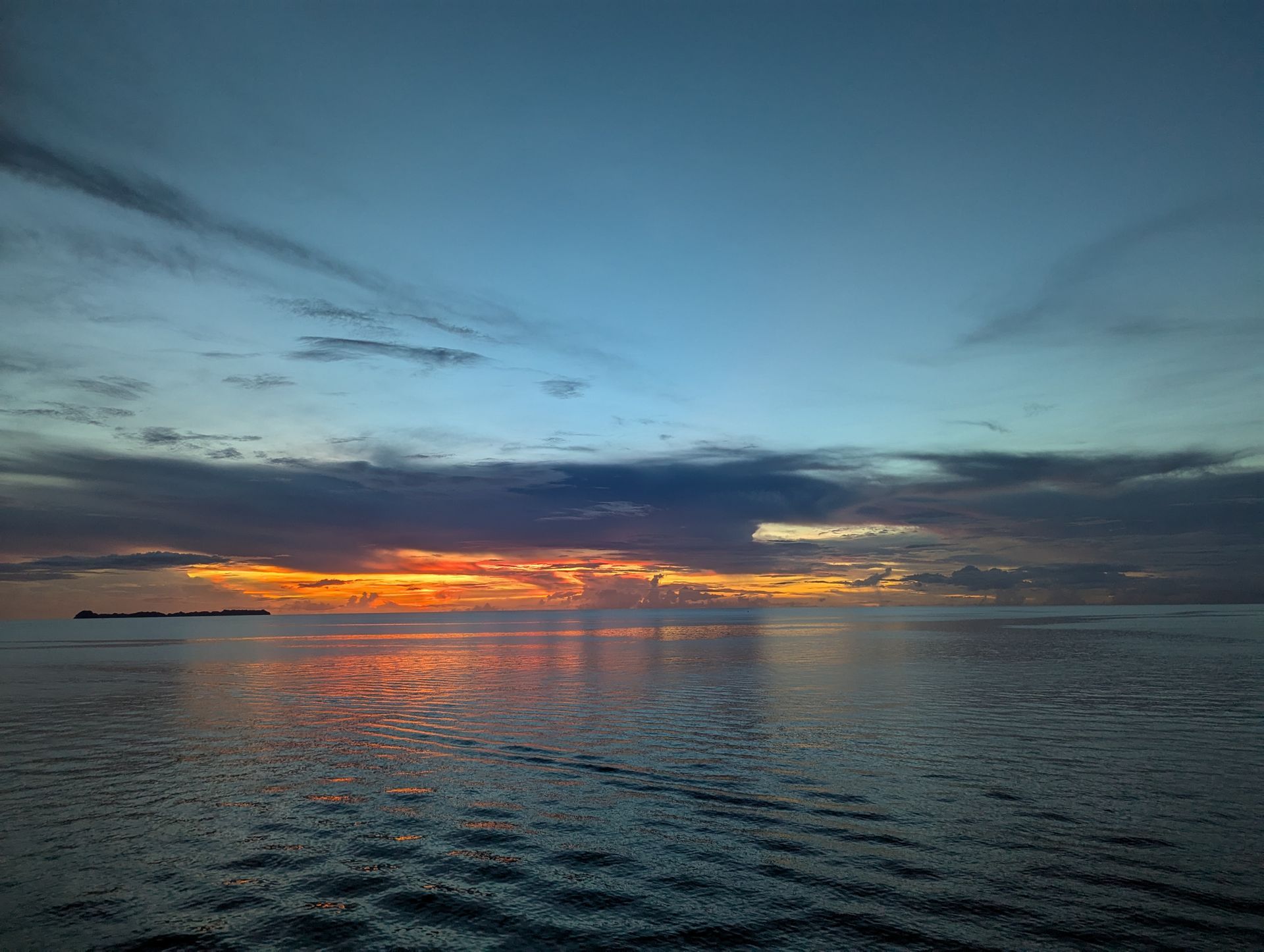 Sunset over ocean; orange and yellow hues reflect on water beneath a cloudy blue sky.