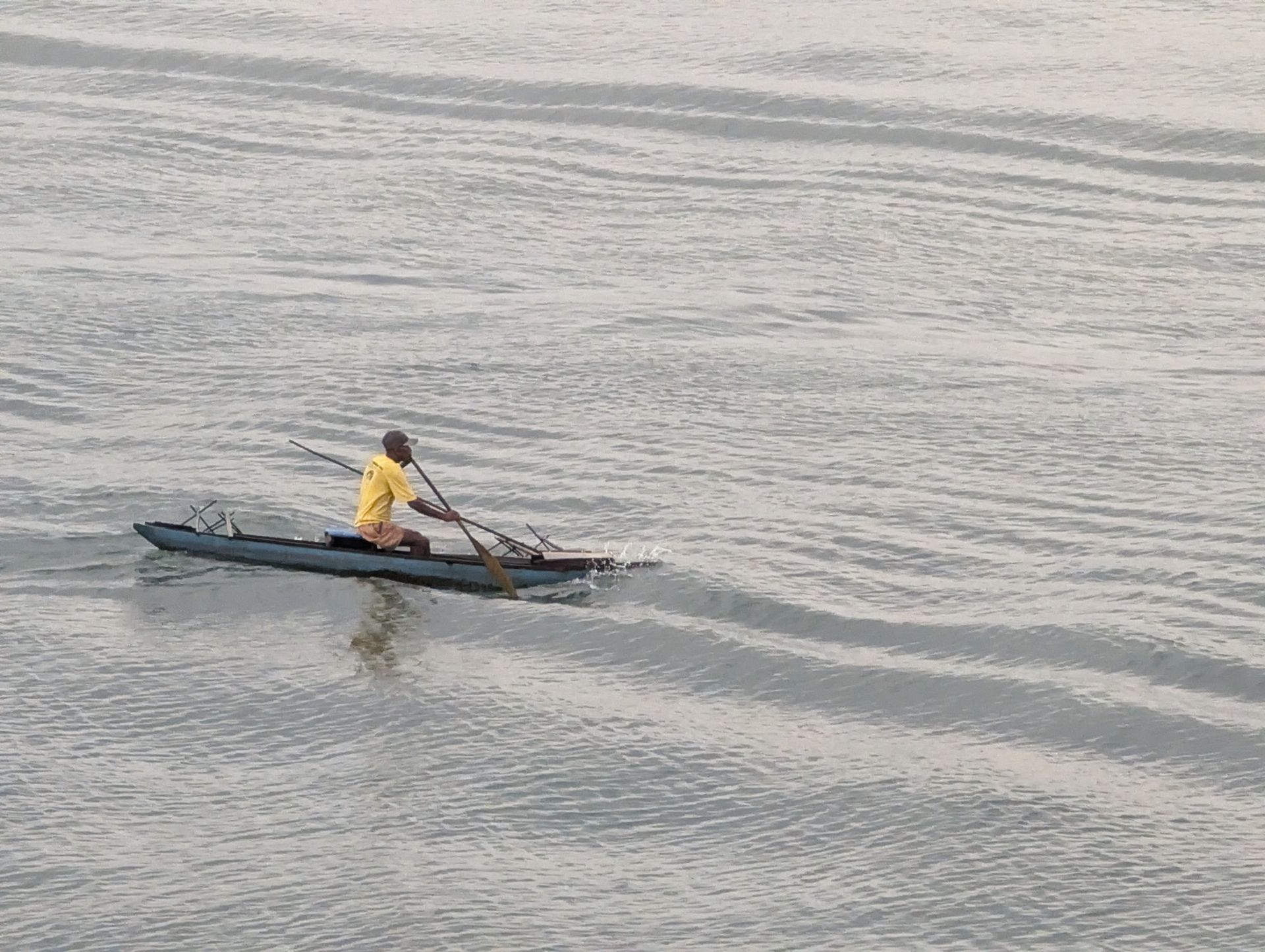 Man in yellow shirt paddles a dark boat on rippled water.