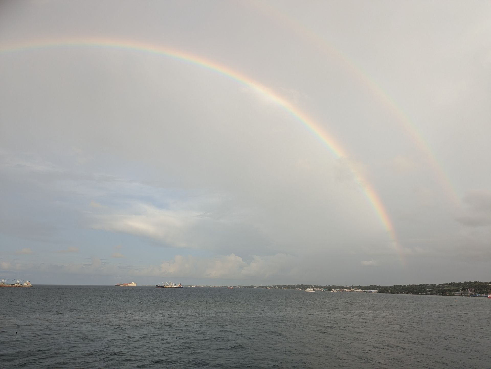 Double rainbow arcs over ocean, ships, and a distant shoreline against a gray sky.