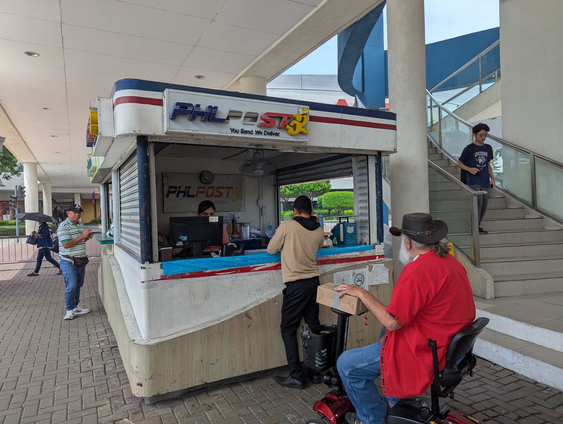 PHLPost kiosk with customers outside, one in a wheelchair. People walk on sidewalk.