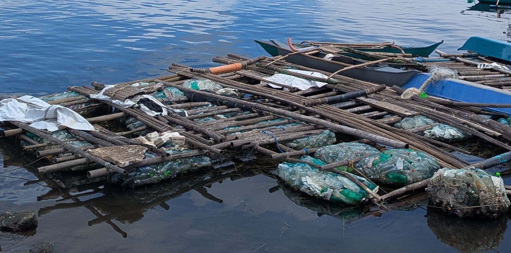 Floating platform made of wood, plastic bottles, and debris in water. Blue boats in the background.