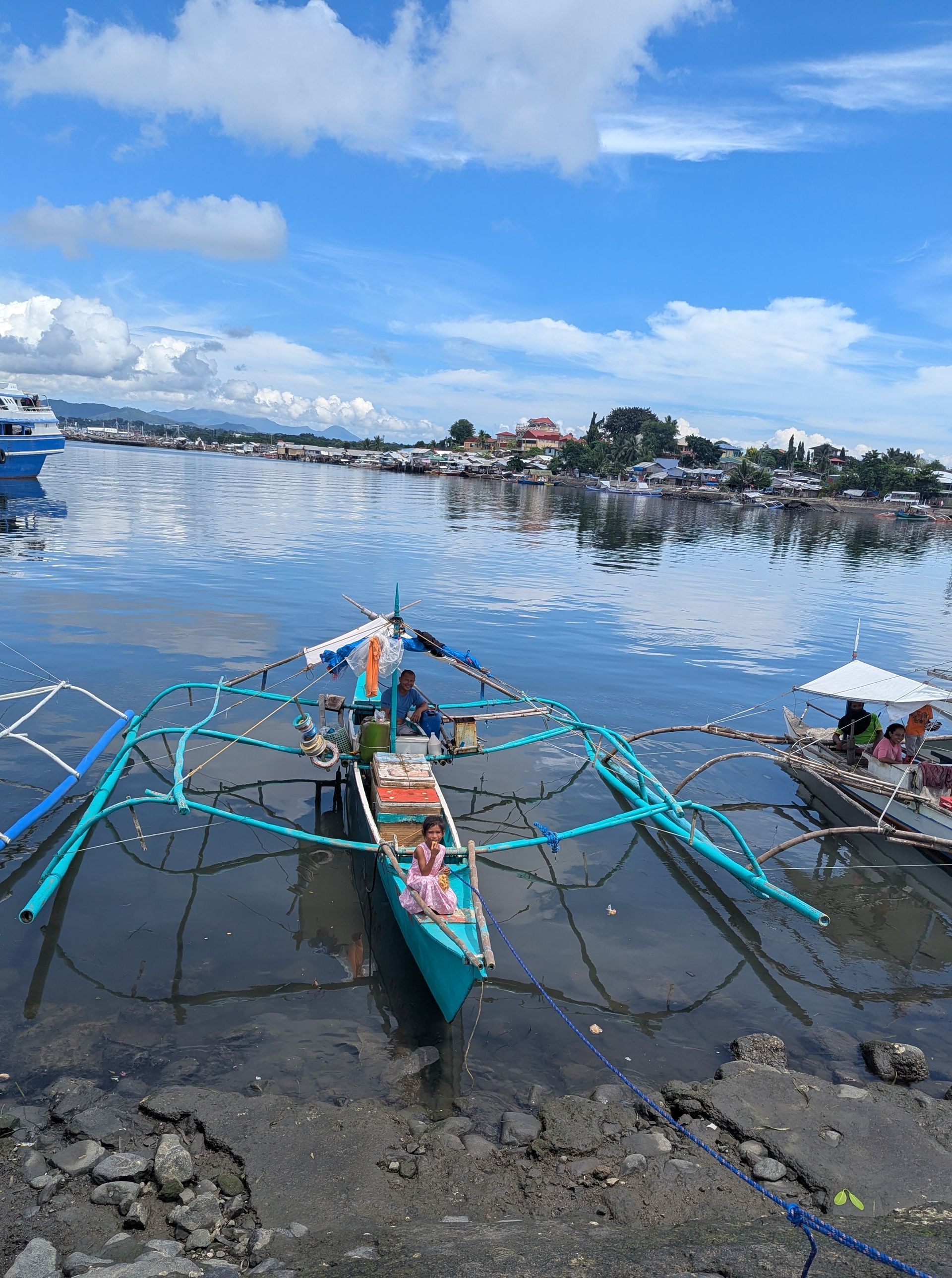 A small turquoise boat in a harbor, with a town on the horizon under a blue sky.