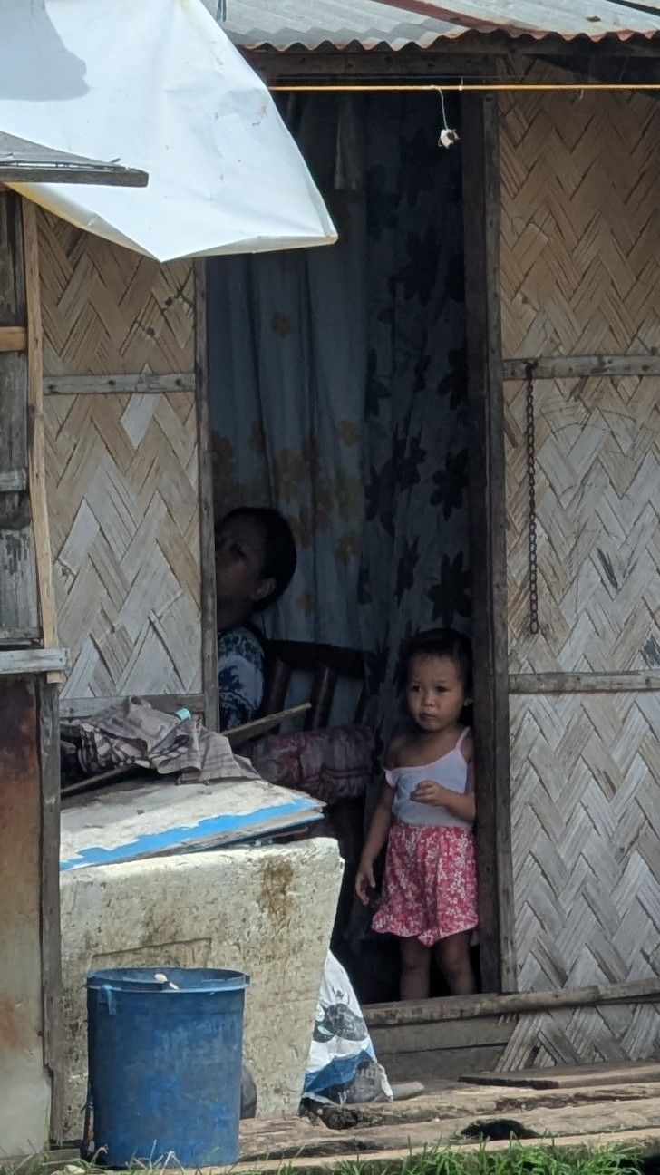 Two children inside a small dwelling. One stands in the doorway wearing pink shorts, the other sits.
