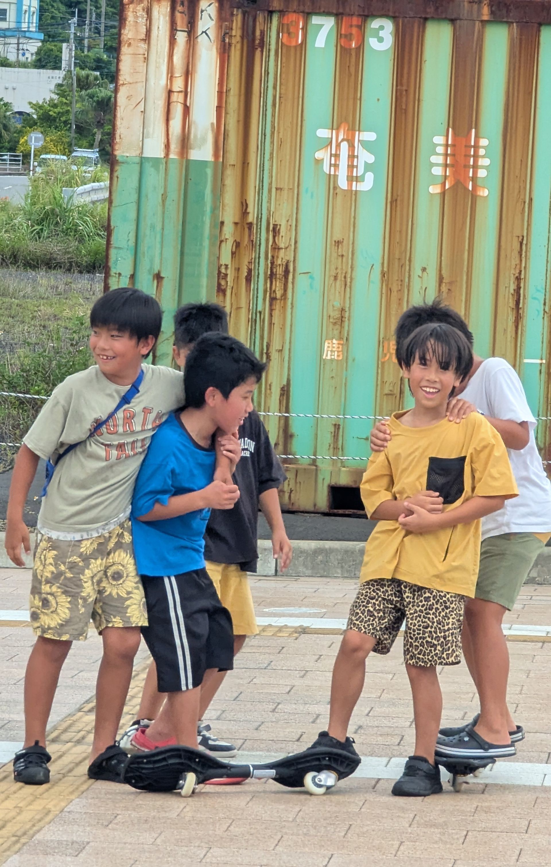 Group of kids on skateboards, laughing and smiling, near a rusty green wall.