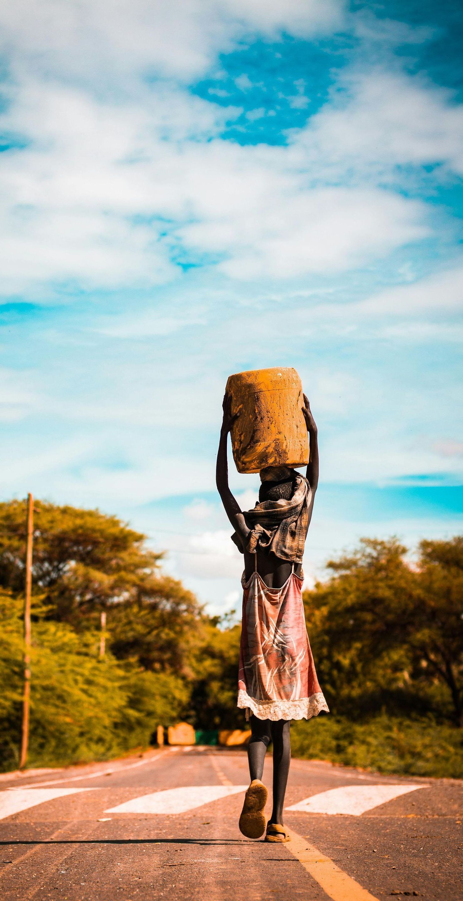 A woman is walking down a road carrying a bucket on her head.