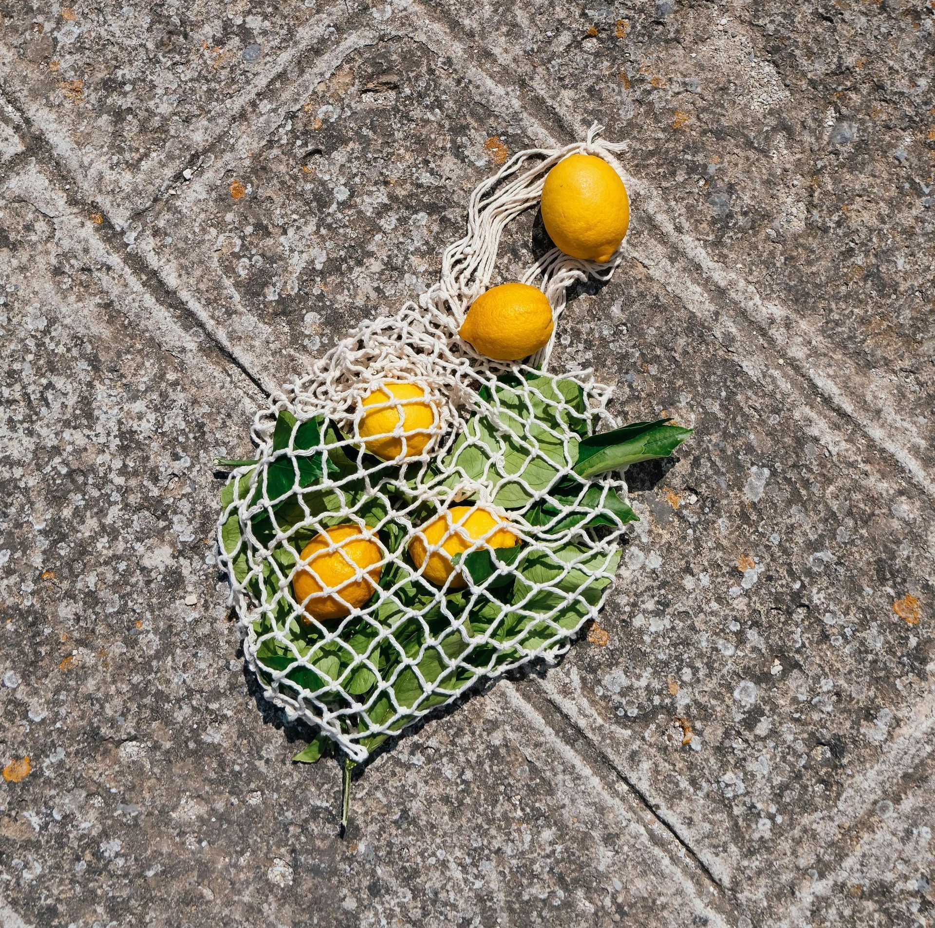 A net bag filled with lemons and green leaves is laying on the ground.