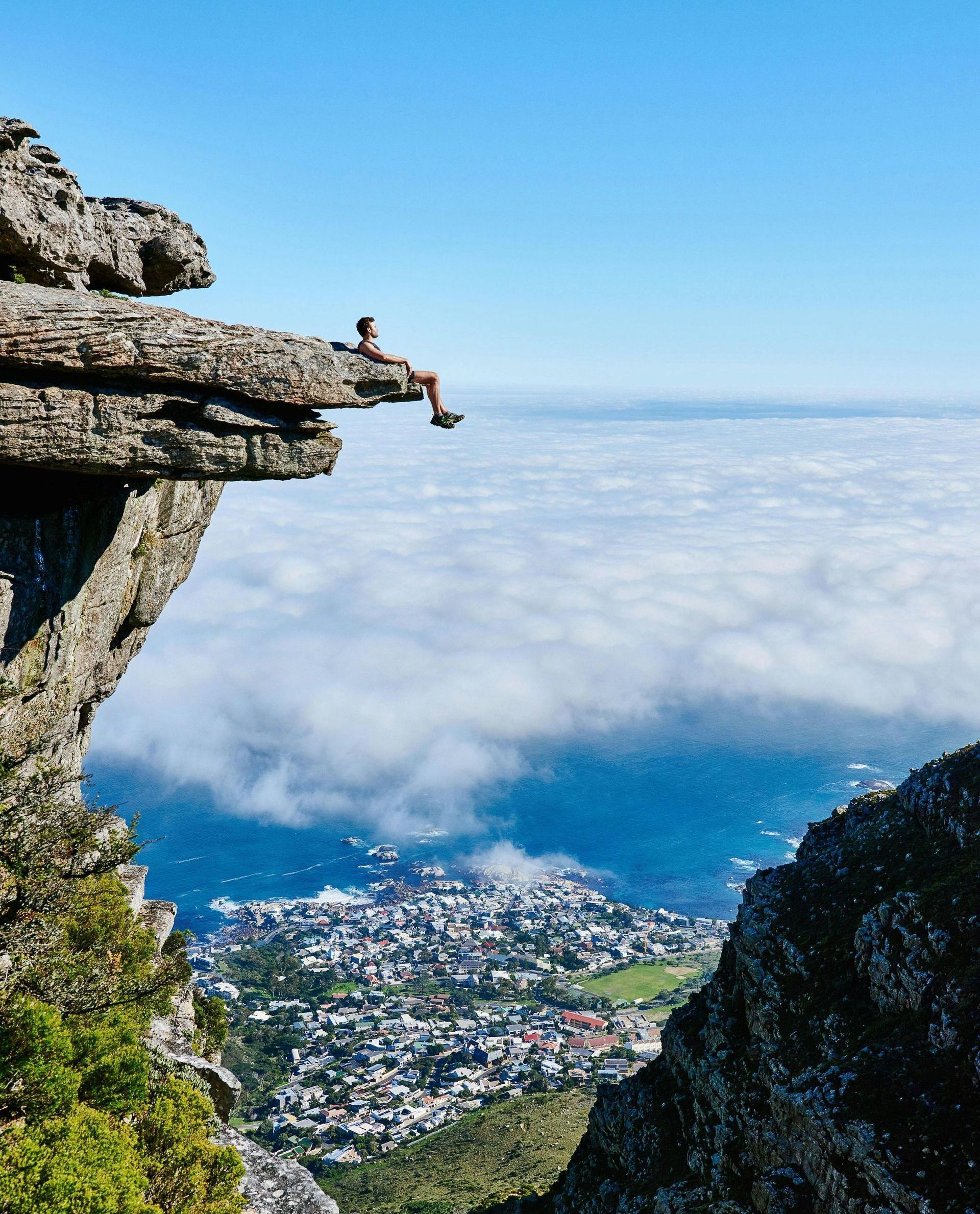 A man is sitting on the edge of a cliff overlooking a city.