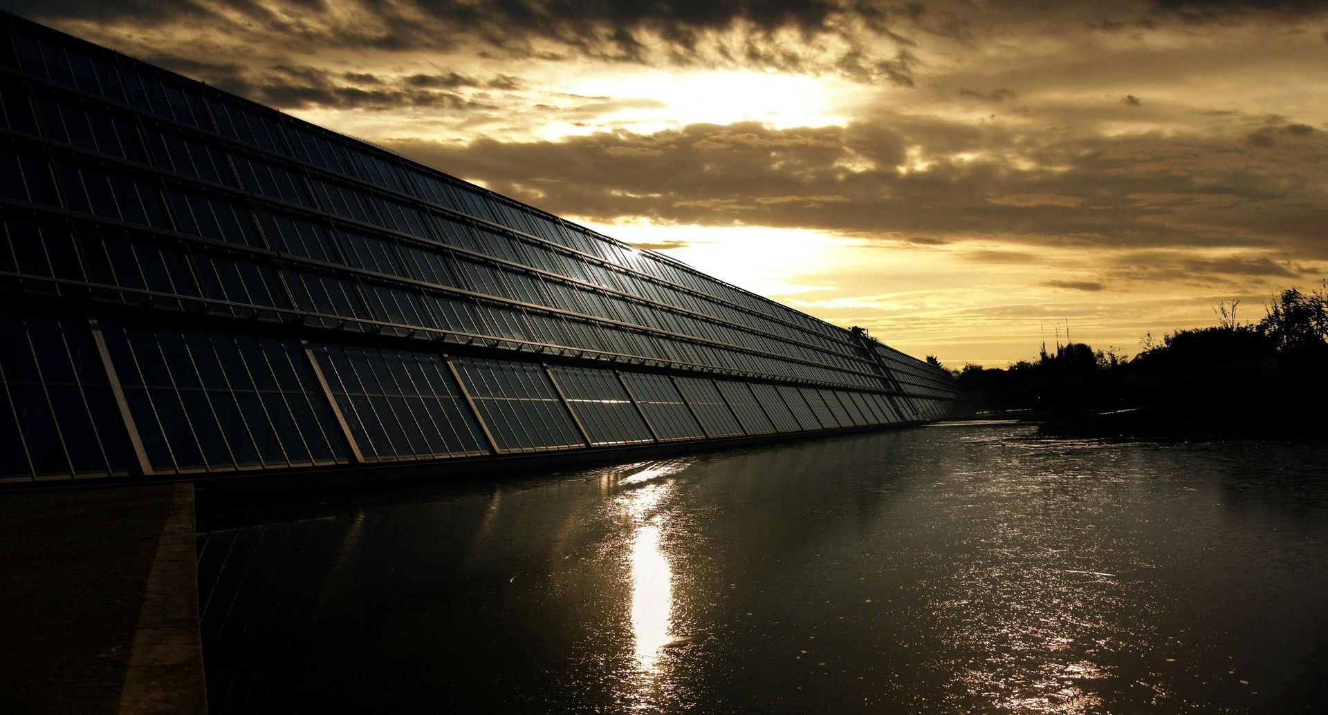 A row of solar panels sitting next to a body of water at sunset.