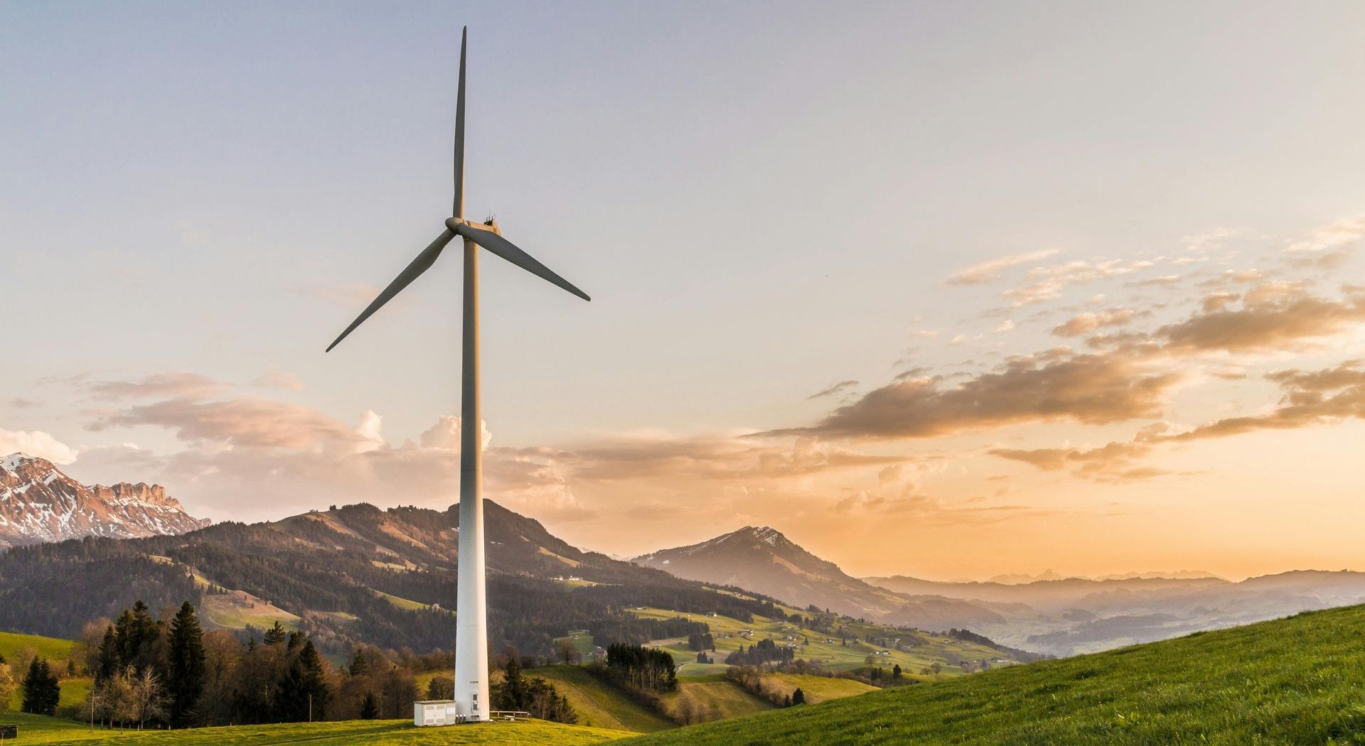 A wind turbine is sitting on top of a grassy hill with mountains in the background.