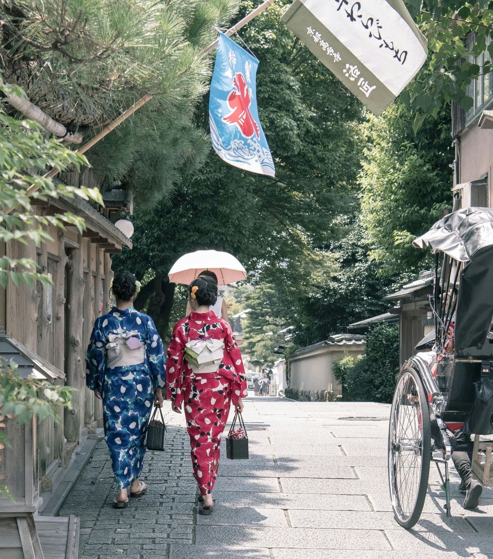 Two women in kimonos are walking down a sidewalk next to a rickshaw.