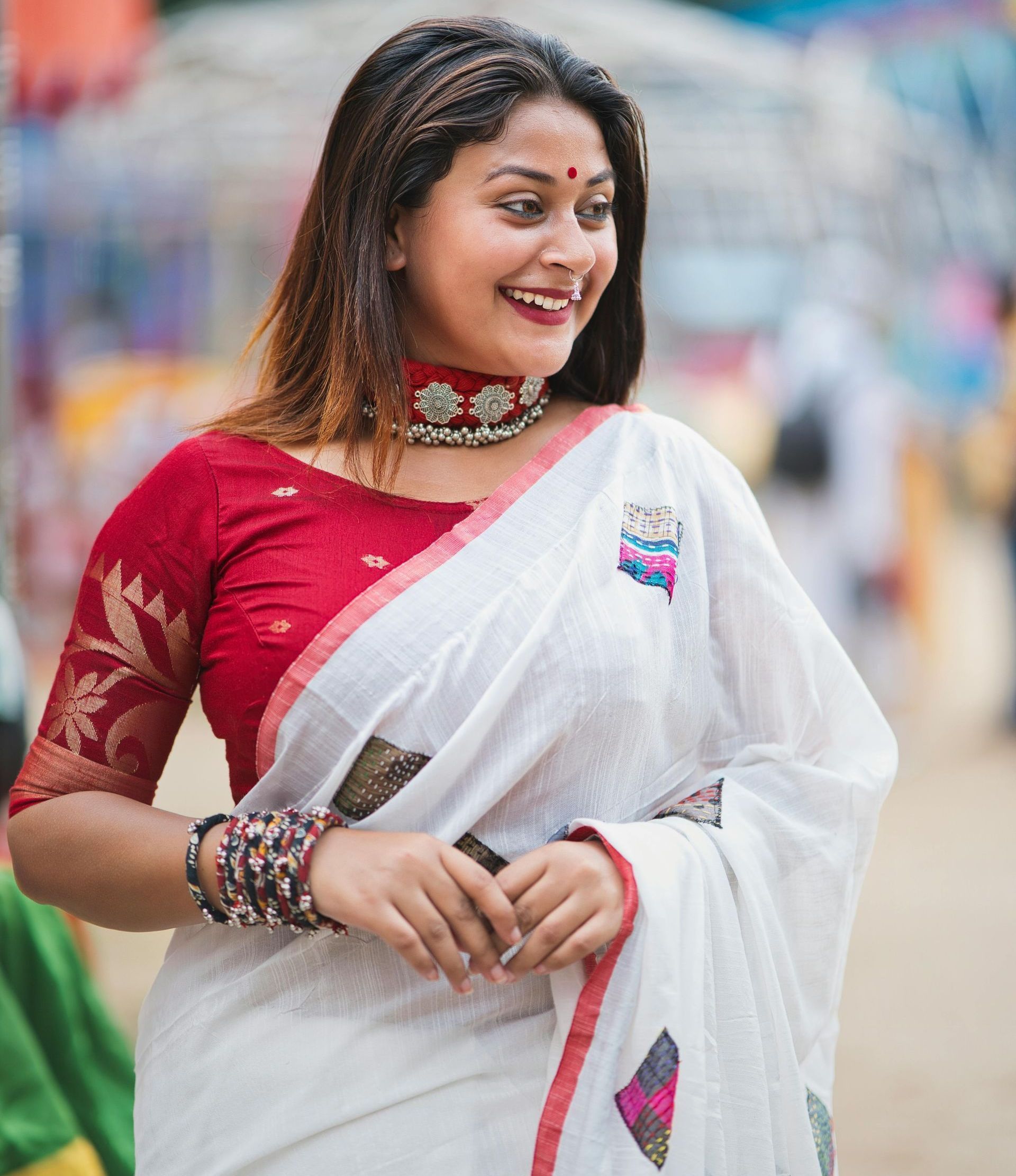 A woman wearing a white saree and a red blouse is smiling.