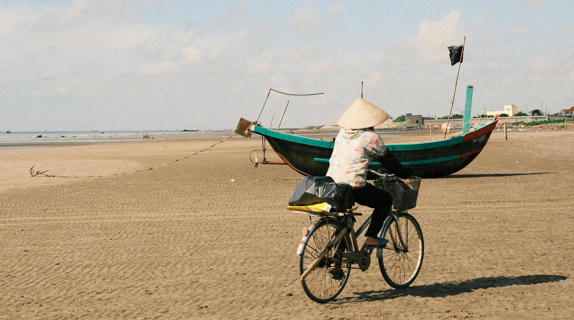 A woman wearing a conical hat is riding a bike on the beach