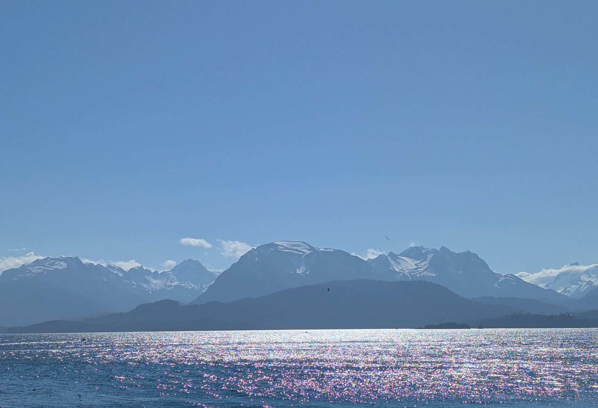 Bright blue sky over sparkling water, with silhouetted mountains in the distance, some snow-capped.