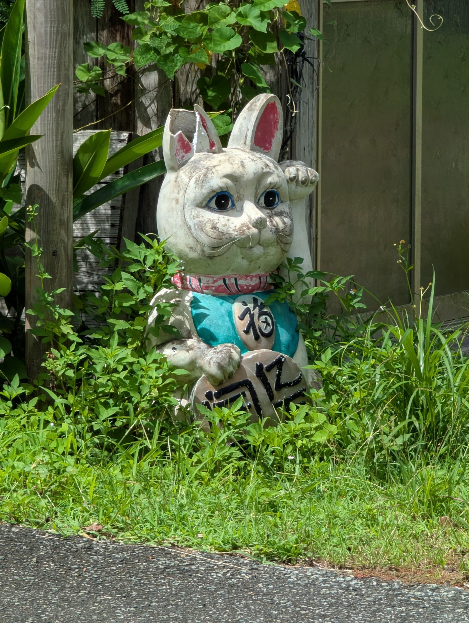 A weathered white rabbit statue with a raised paw, wearing a teal bib, surrounded by greenery.