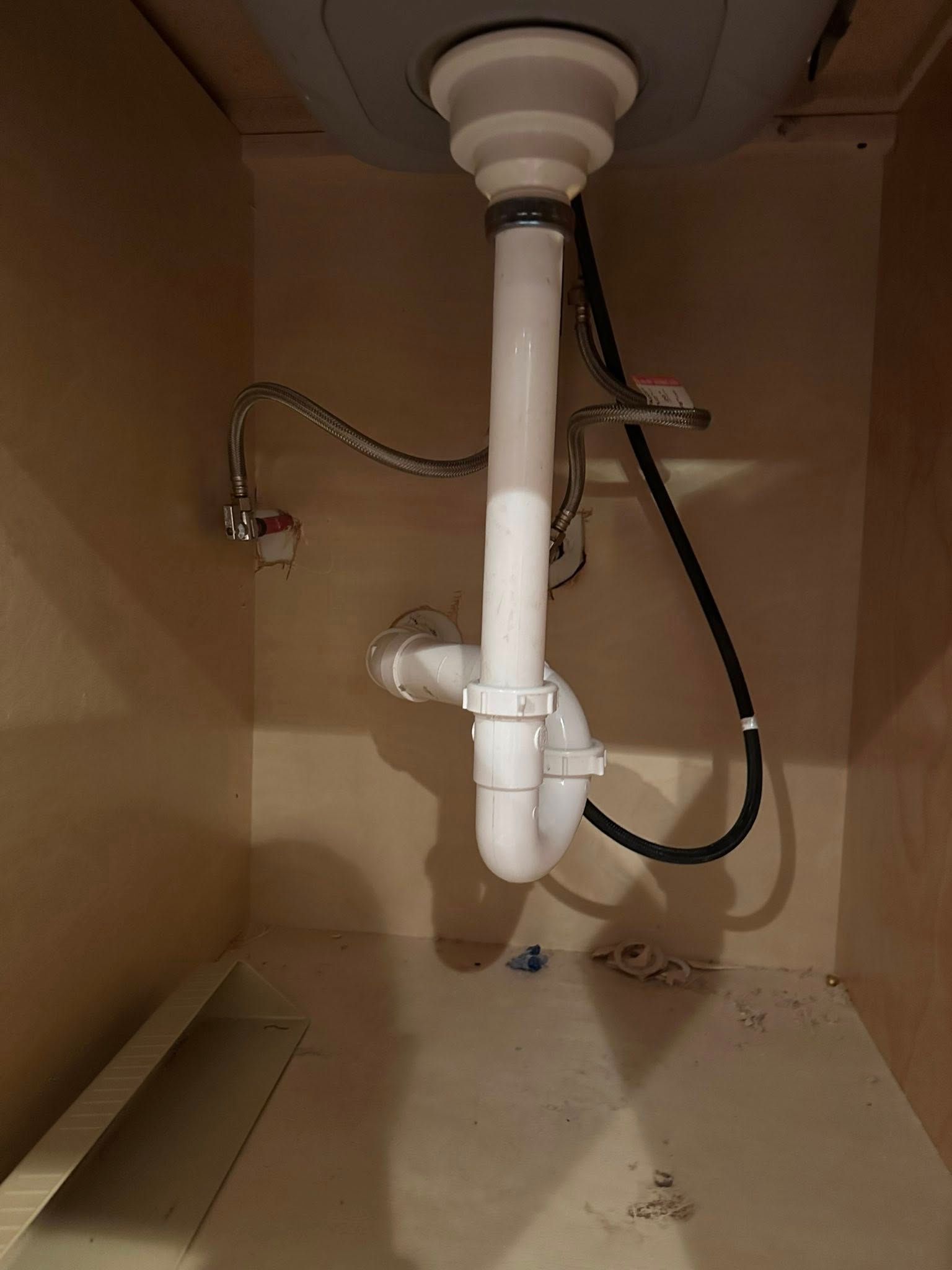 White plumbing pipes under a sink in a beige cabinet. Silver faucet pipe and black electrical wire are visible.