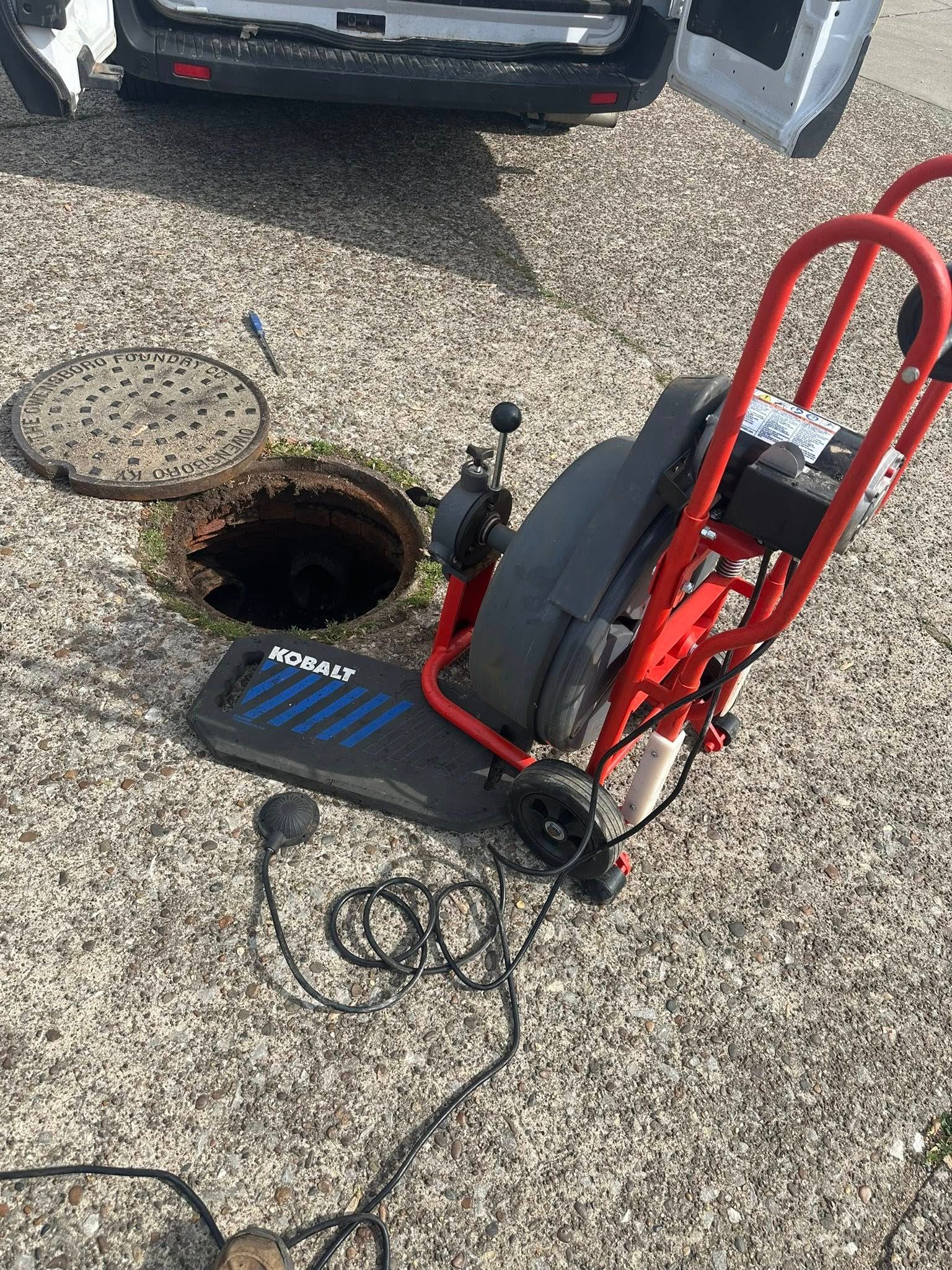 Plumber's drain cleaning machine next to an open manhole. Red and black equipment on gravel.