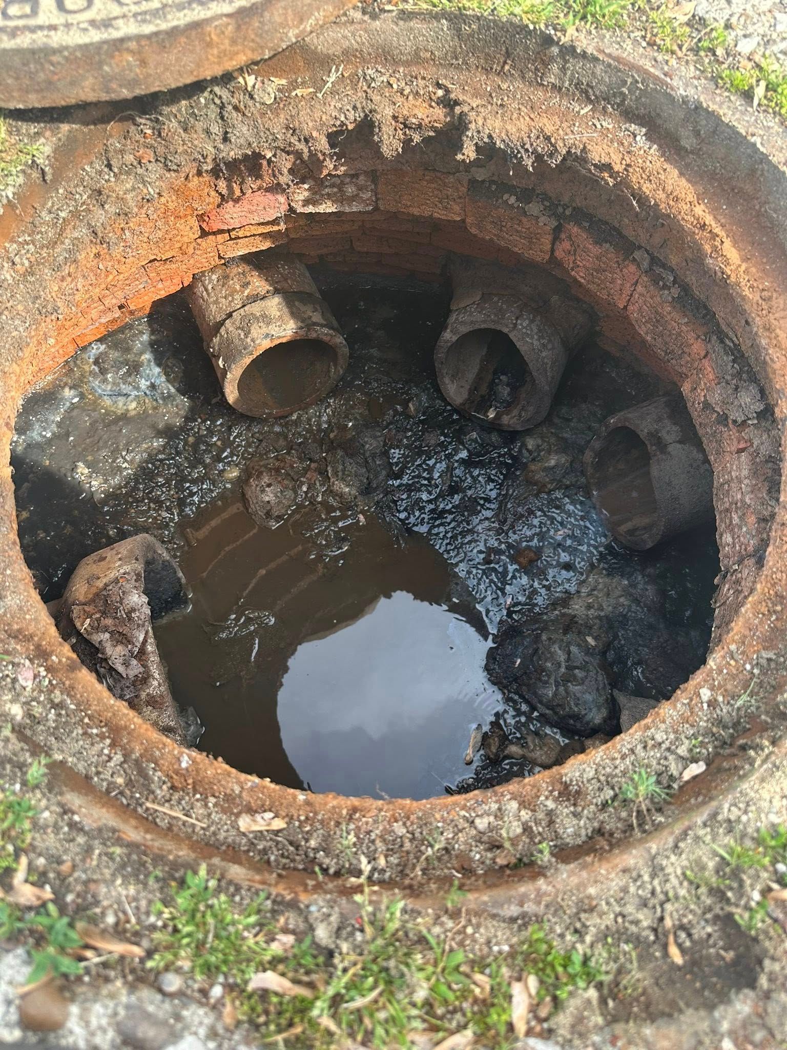 Open manhole with pipes and standing water, surrounded by grass.