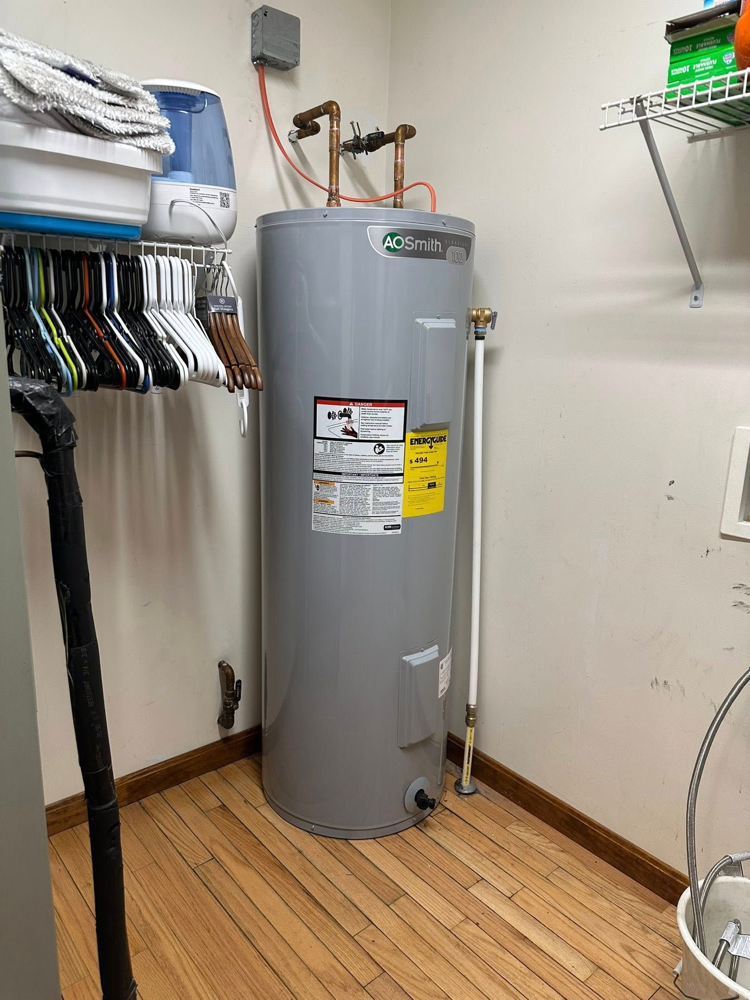 A tall, grey water heater in a laundry room with hardwood floors and a shelf with clothes and accessories.