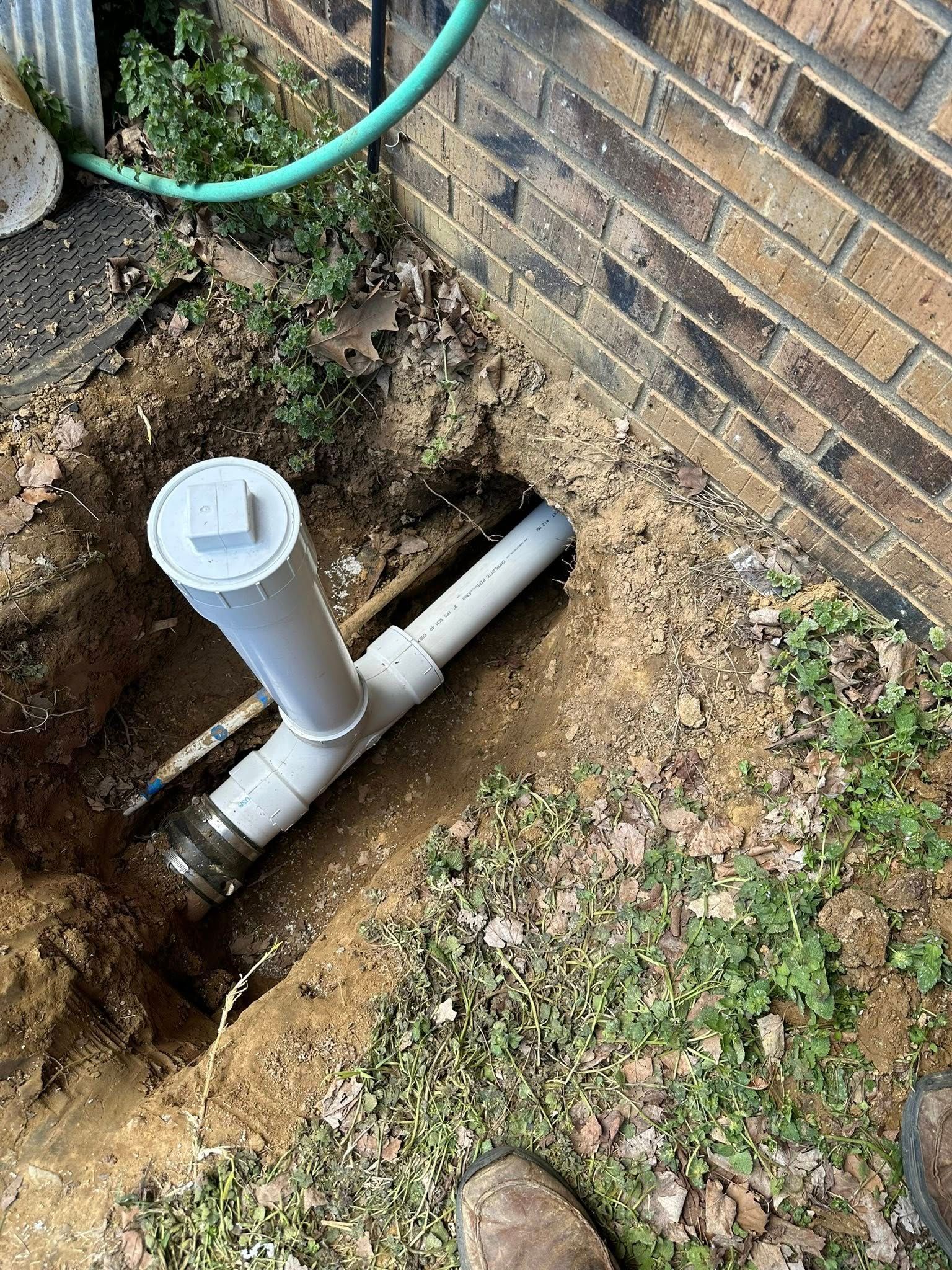 White PVC pipes connected to a brick wall; dug-out trench; green hose; outdoor.