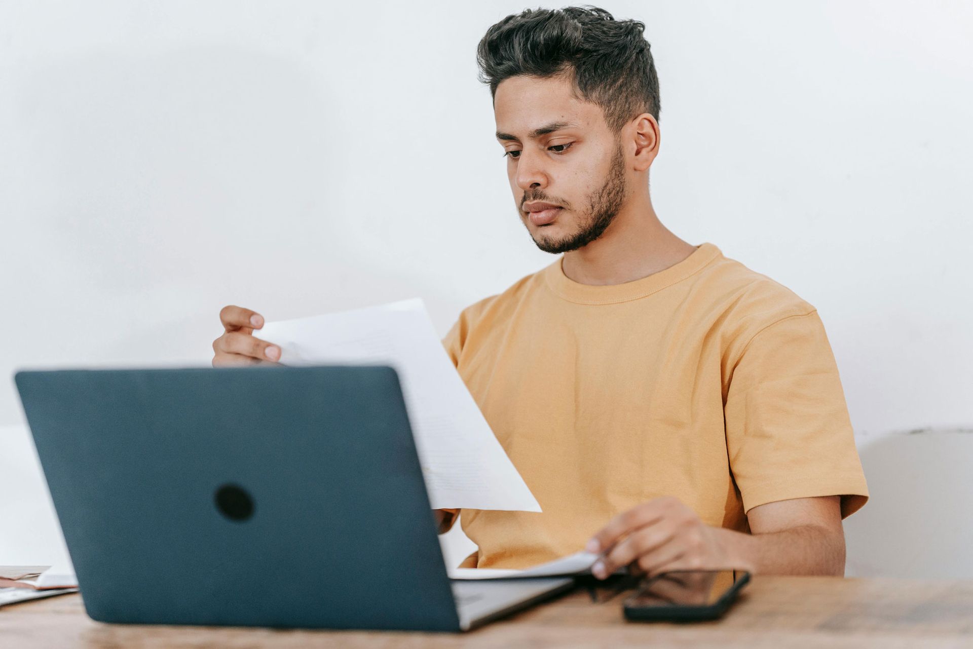 A man is sitting at a table using a laptop computer while holding a piece of paper.