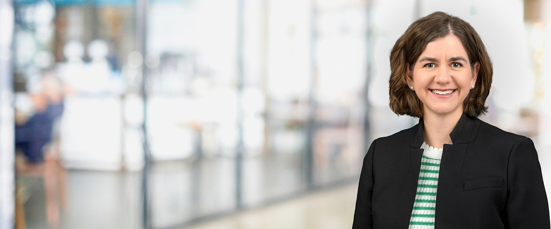 Woman in a black blazer smiles at the viewer in a blurry office setting.