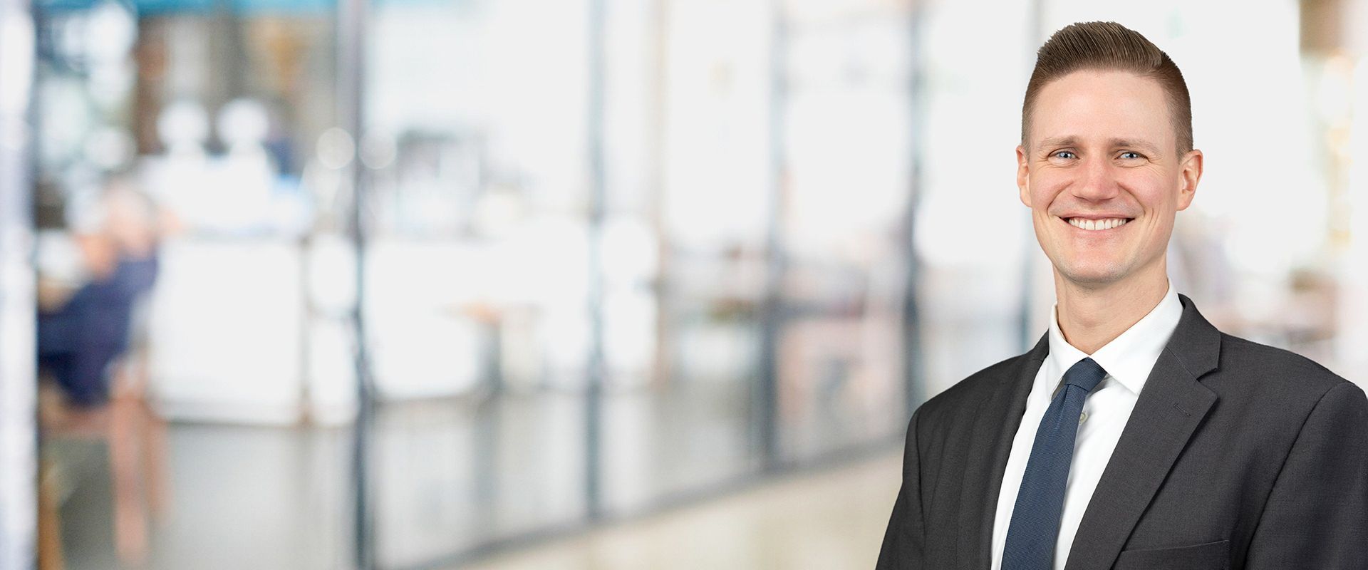 Man in a dark suit and tie, smiling, in front of an office setting.