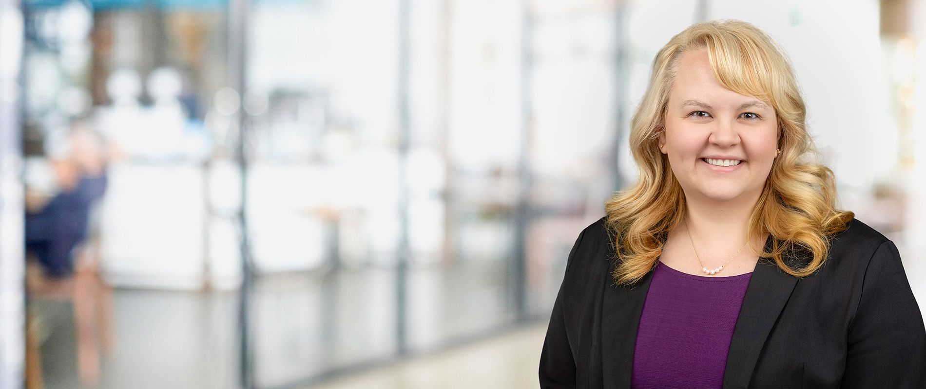 Woman with blonde hair, smiling, in a black blazer and purple top, blurred office background.