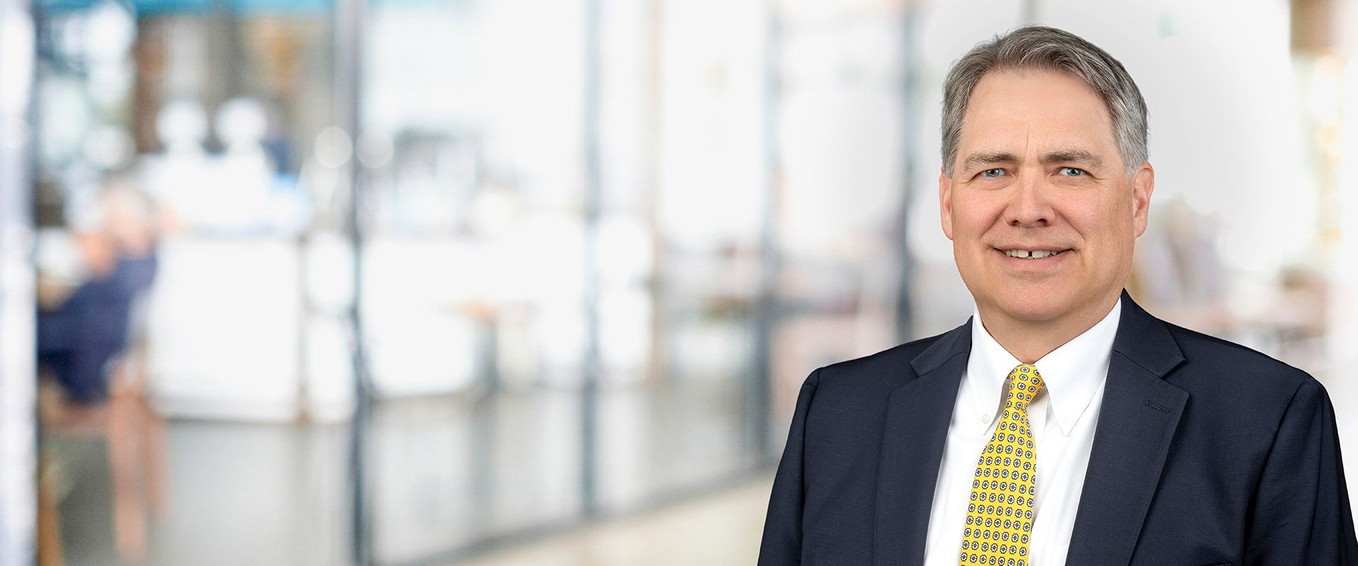 Man in a suit smiles, tie with yellow pattern, blurred office background.