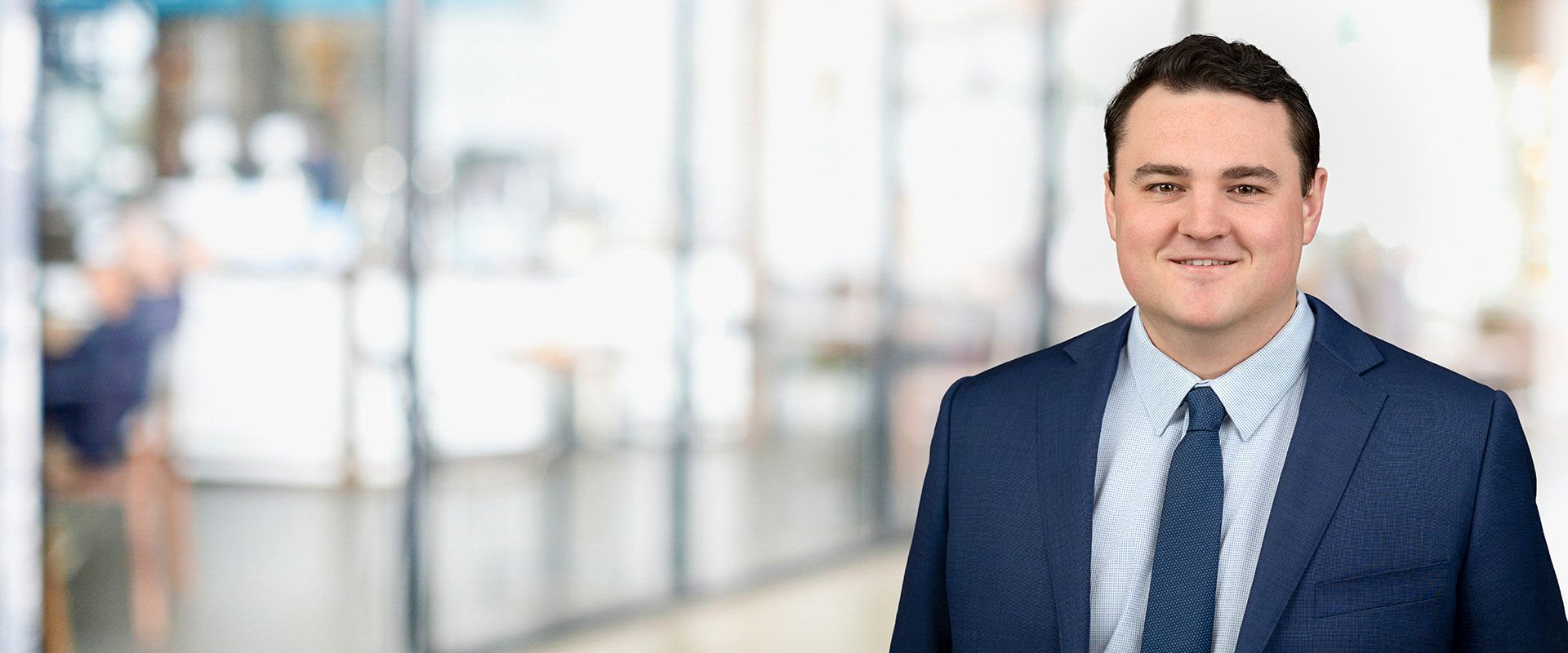 A man in a suit and tie smiling. He's standing in an office with blurred background.