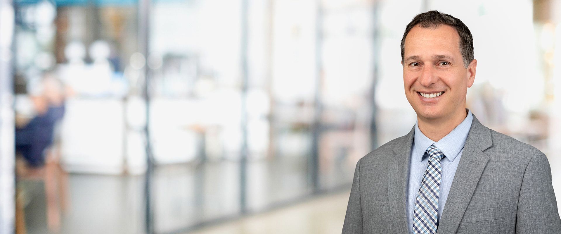 Man in gray suit and tie smiles, blurred background.
