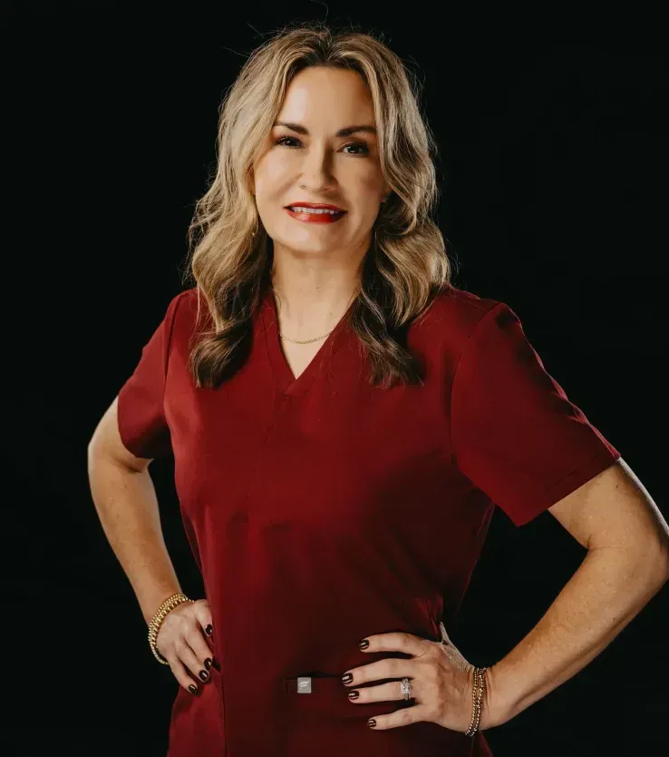 Professional headshot of a person in maroon medical scrubs, posing with hands on hips against a black background.